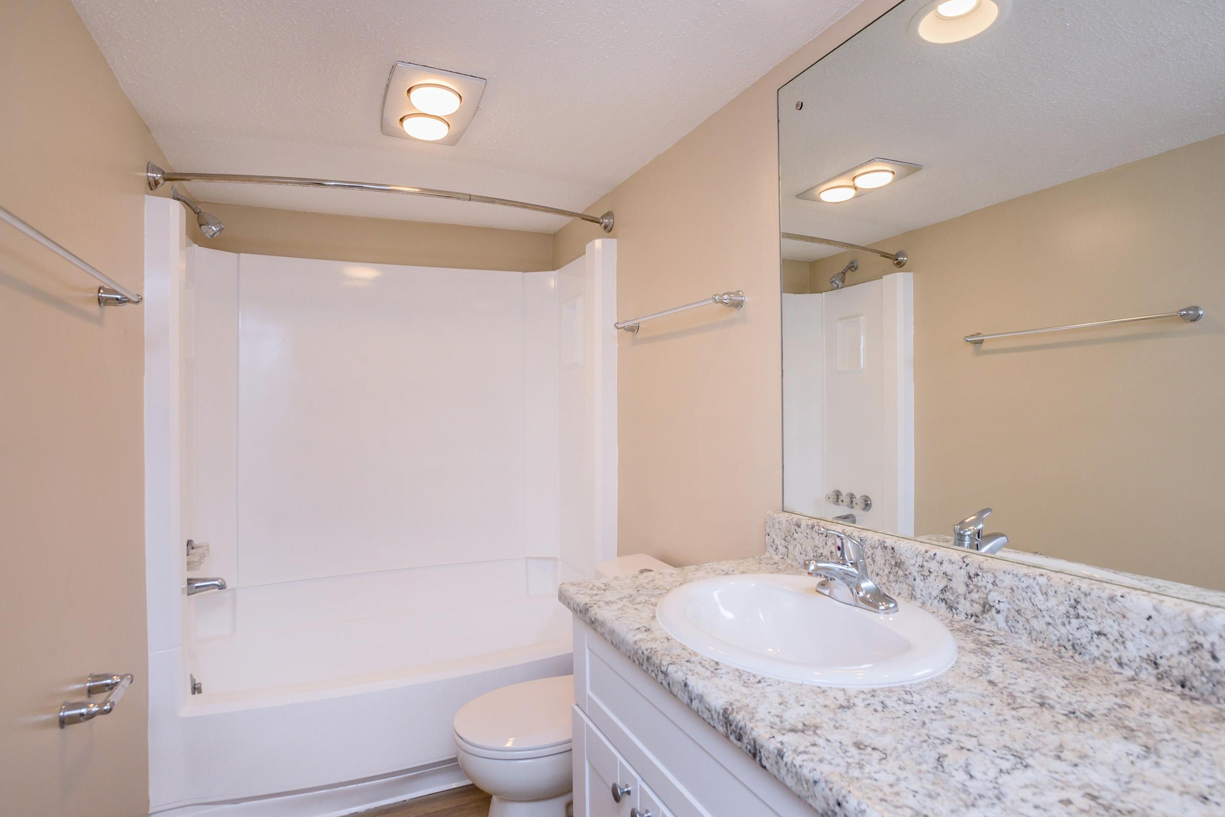 A clean and modern bathroom featuring a white bathtub and shower combination, a white toilet, and a sink with a granite countertop. The walls are painted a light beige, and there is a large mirror above the sink. Bright overhead lighting enhances the space.