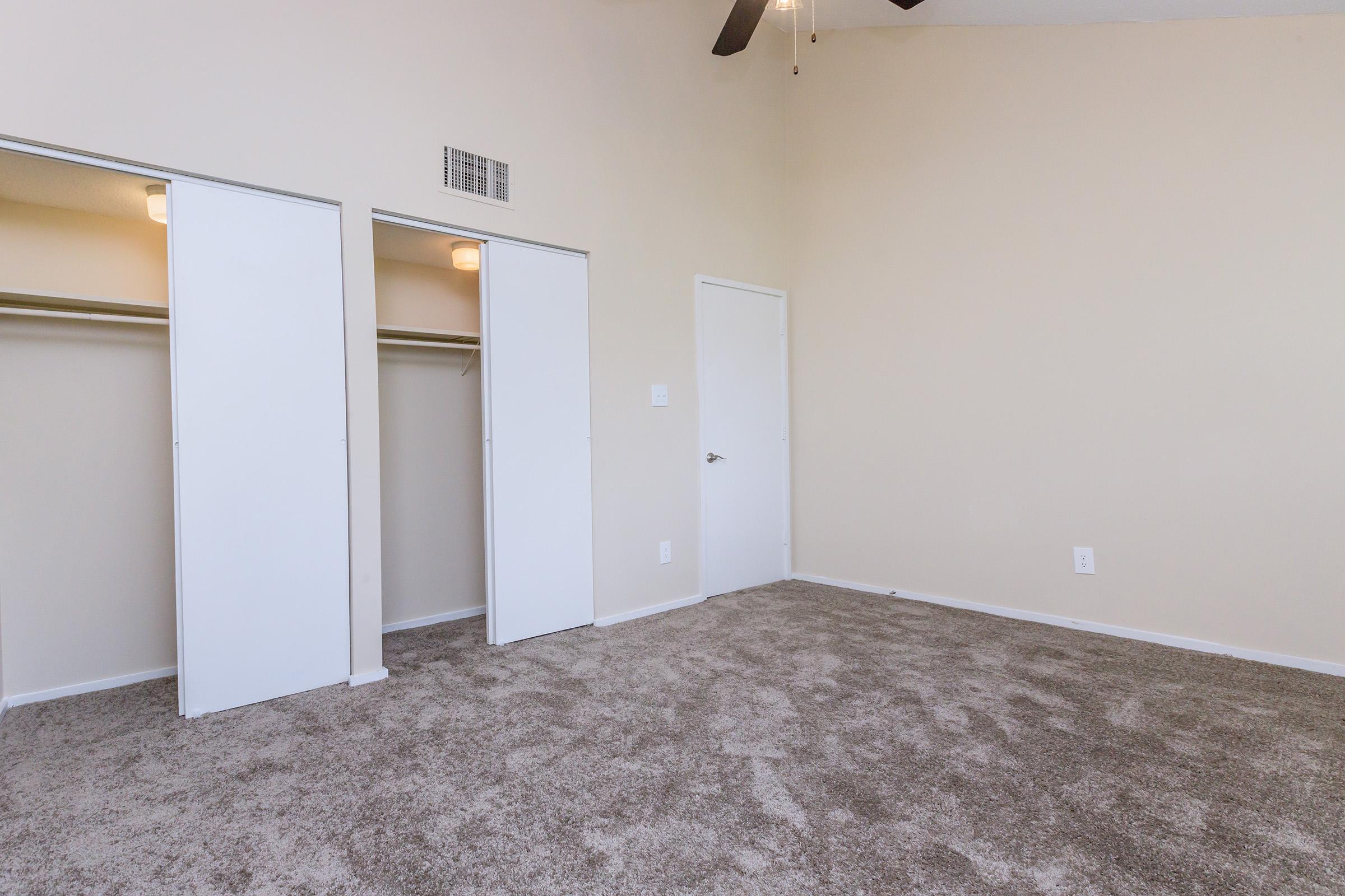Empty room featuring light-colored walls, a ceiling fan, and plush carpeting. Two white sliding closet doors are visible on the left side, and a single door is located on the right. Natural light illuminates the space, creating a bright, airy atmosphere.