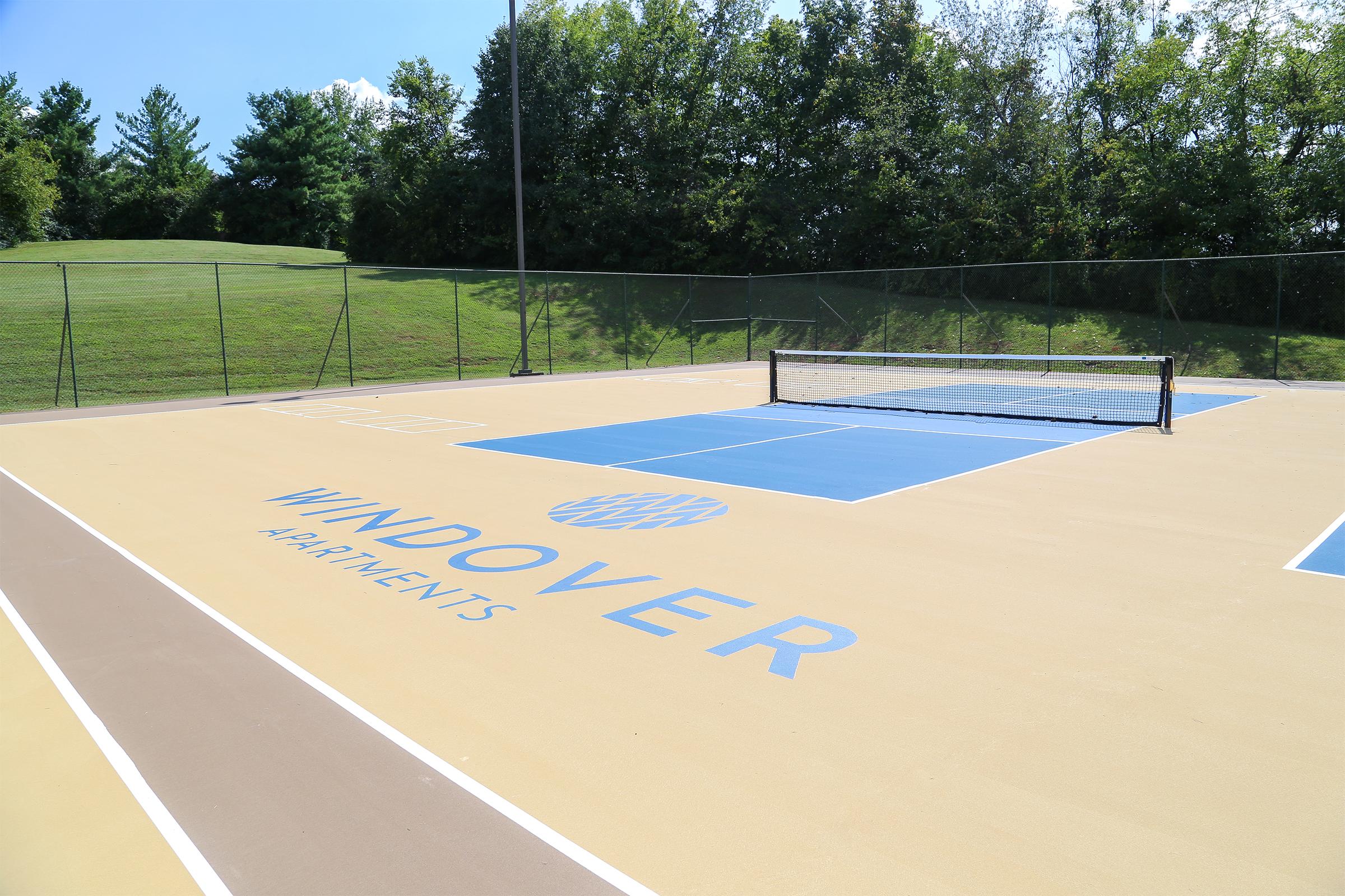 A tennis court with a light tan surface featuring blue accents. The court is marked with the name "WINDOVER APARTMENTS" in large letters. Surrounding the court are green trees and grass, under a clear blue sky. There is a tennis net in the middle of the court.