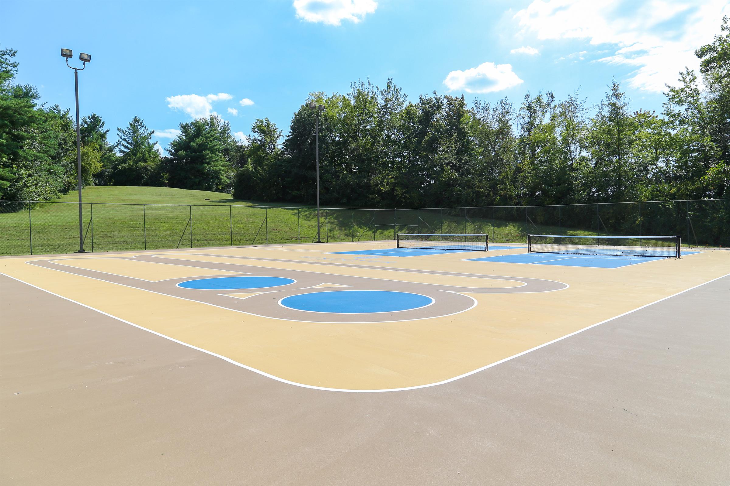 A well-maintained outdoor sports court featuring two blue basketball hoops and colorful markings on a tan surface. The background includes green trees and a blue sky with fluffy clouds, creating a bright and inviting atmosphere for recreational activities.