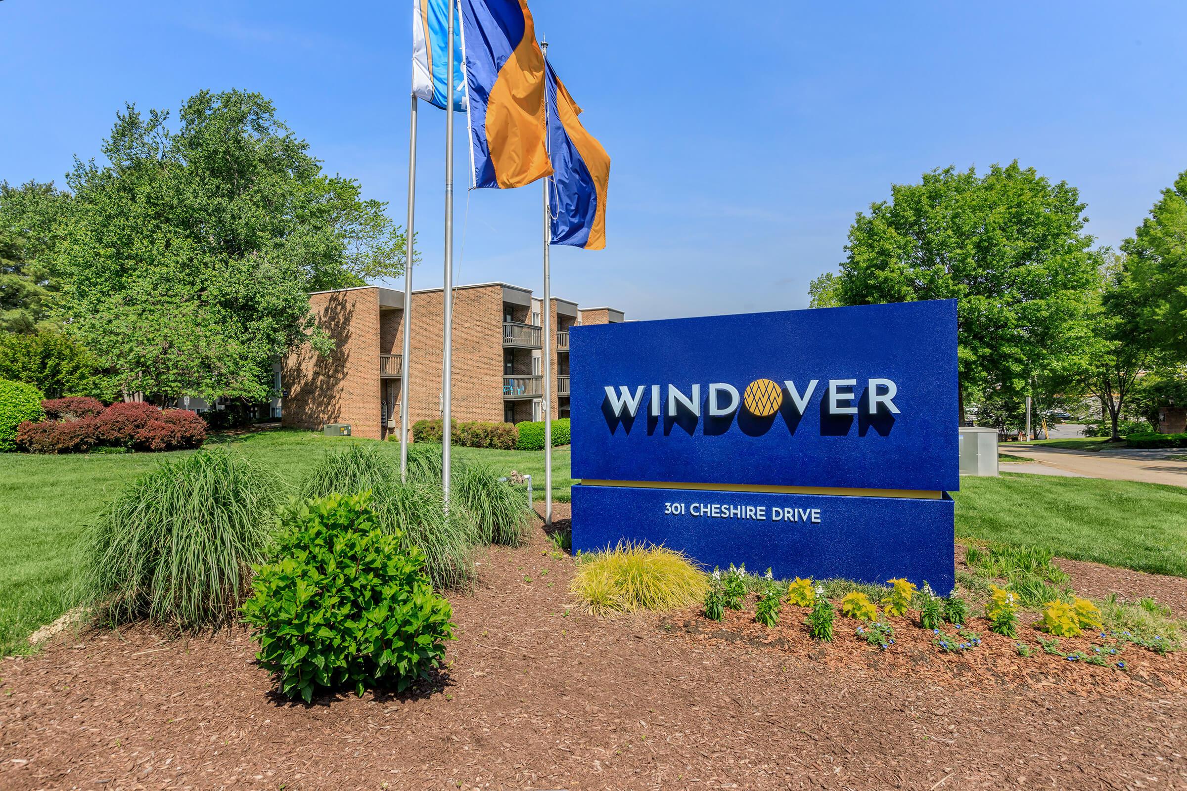 A blue sign displaying the name "WINDOVER" and the address "301 CHESHIRE DRIVE" is surrounded by vibrant green grass and landscaping. There are several flags flying in front of a brick building and trees in the background, set against a clear blue sky.