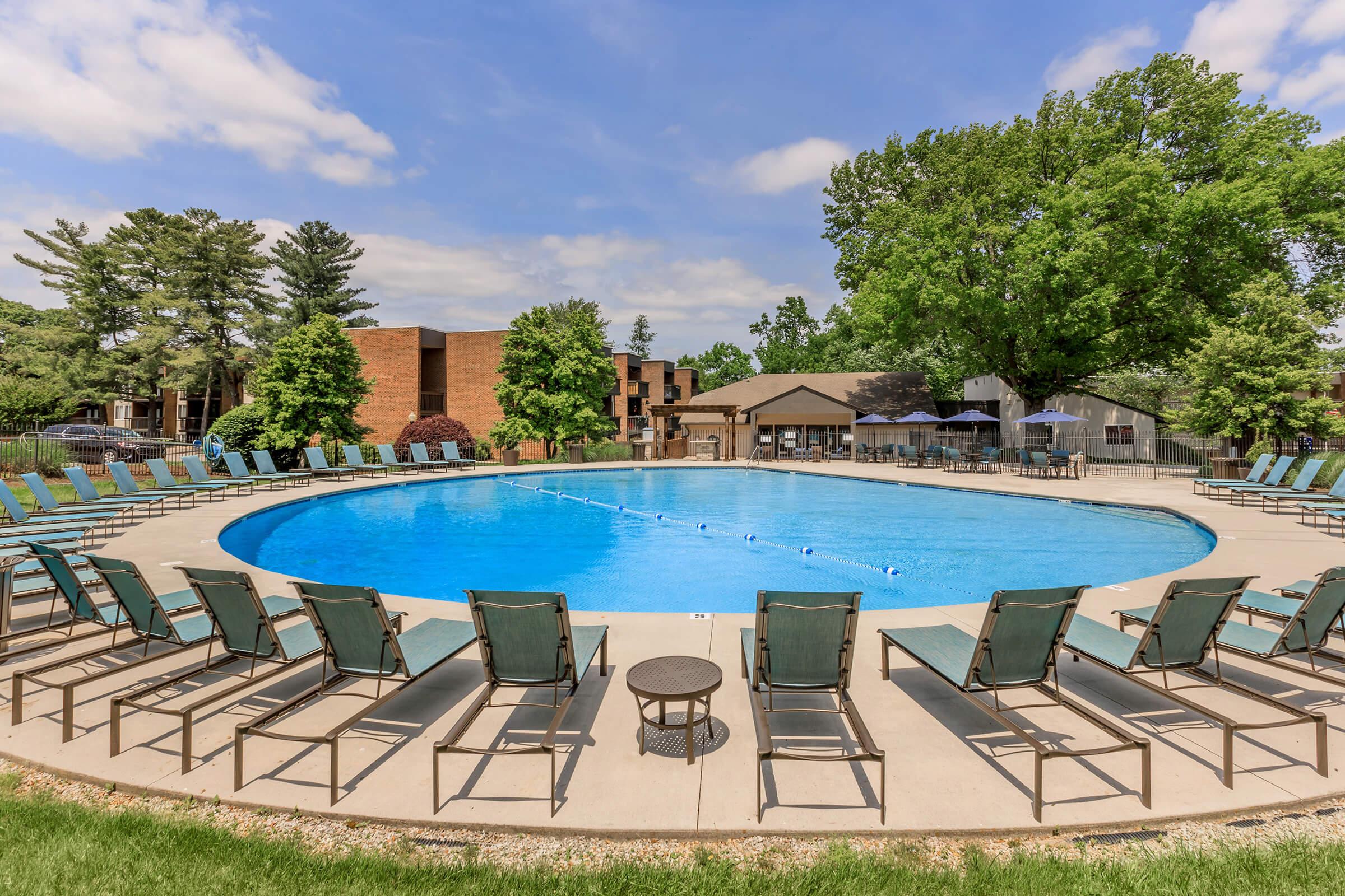 A large outdoor swimming pool surrounded by lounge chairs and lush greenery. Several trees and buildings in the background, with a calm sky above. The scene is designed for relaxation and leisure, perfect for enjoying a sunny day by the water.
