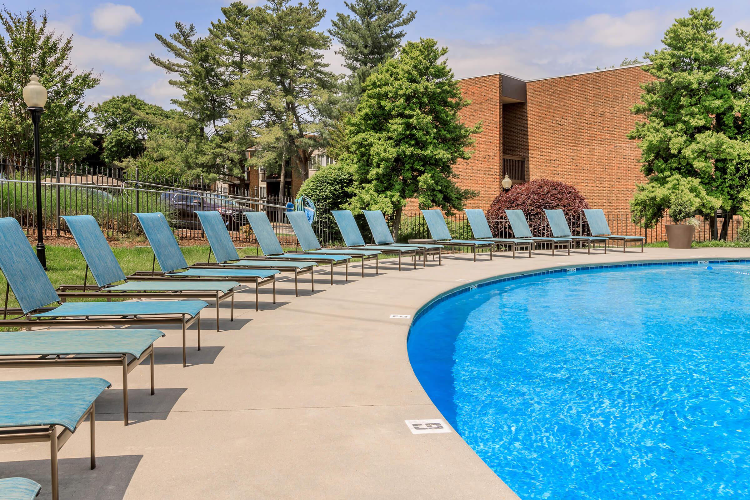 A swimming pool surrounded by empty lounge chairs arranged neatly along the edge. Lush greenery and trees are visible in the background, offering a serene outdoor setting. The sky is partly cloudy, complementing the inviting blue water of the pool.