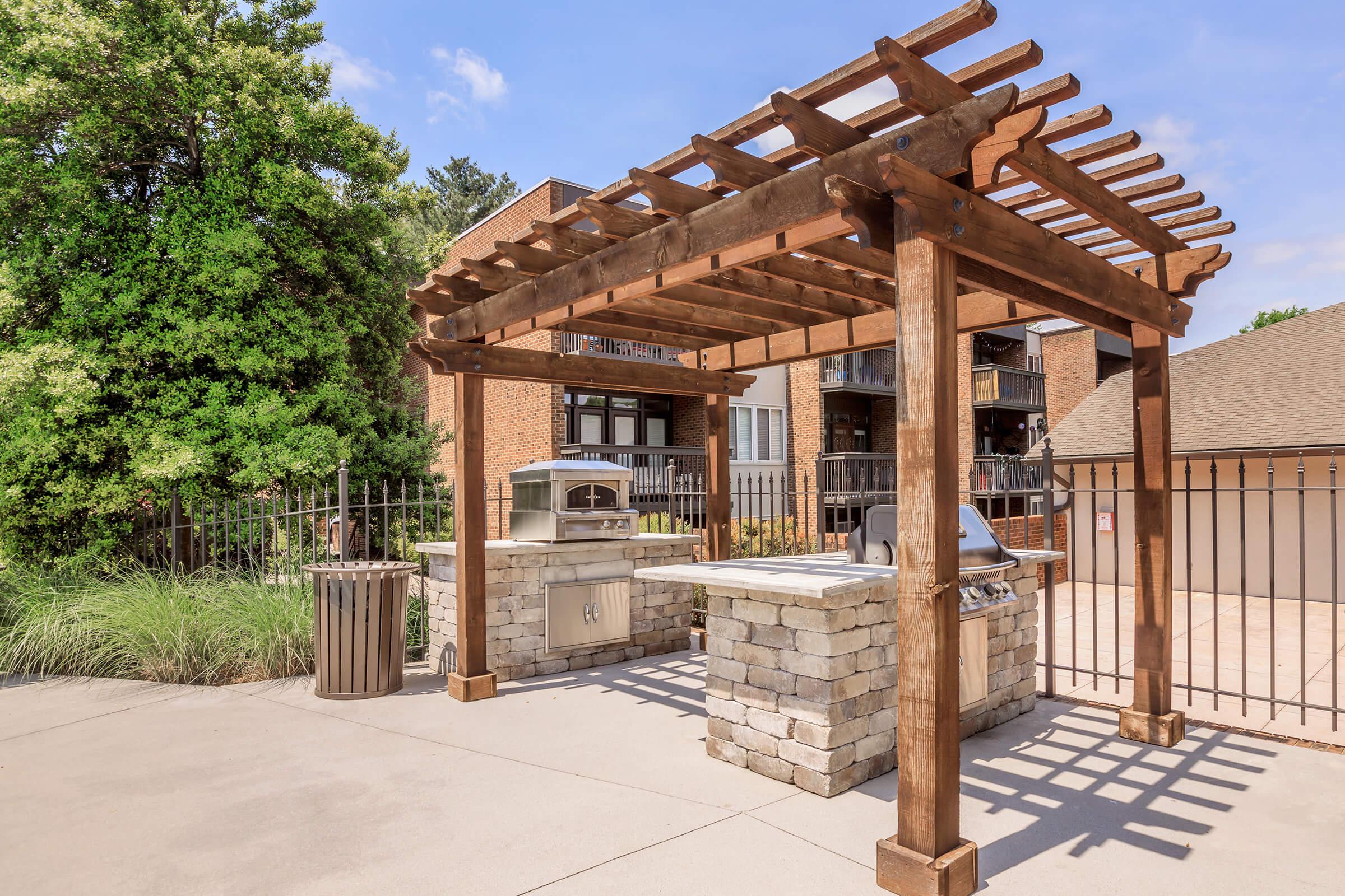 Outdoor grilling area featuring a stone grill station with a wooden pergola overhead, surrounded by a fence and greenery, against a backdrop of apartment buildings and a clear blue sky.