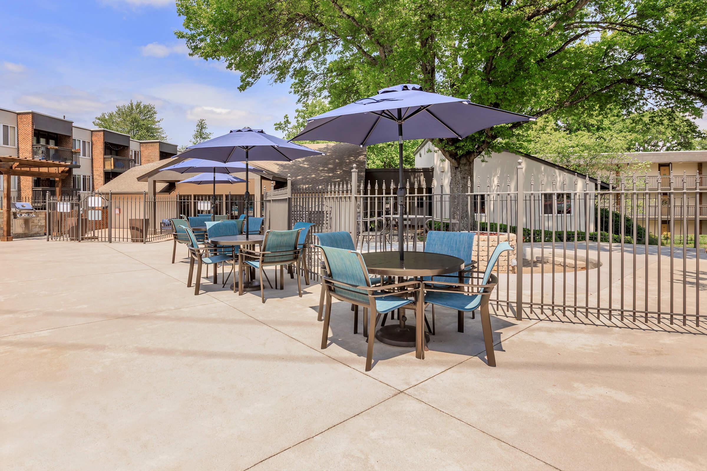 Outdoor seating area featuring several round tables with blue chairs and large umbrellas, set against a backdrop of trees and nearby buildings. The space is paved and enclosed by a fence, creating a comfortable and inviting atmosphere for relaxation or outdoor dining.