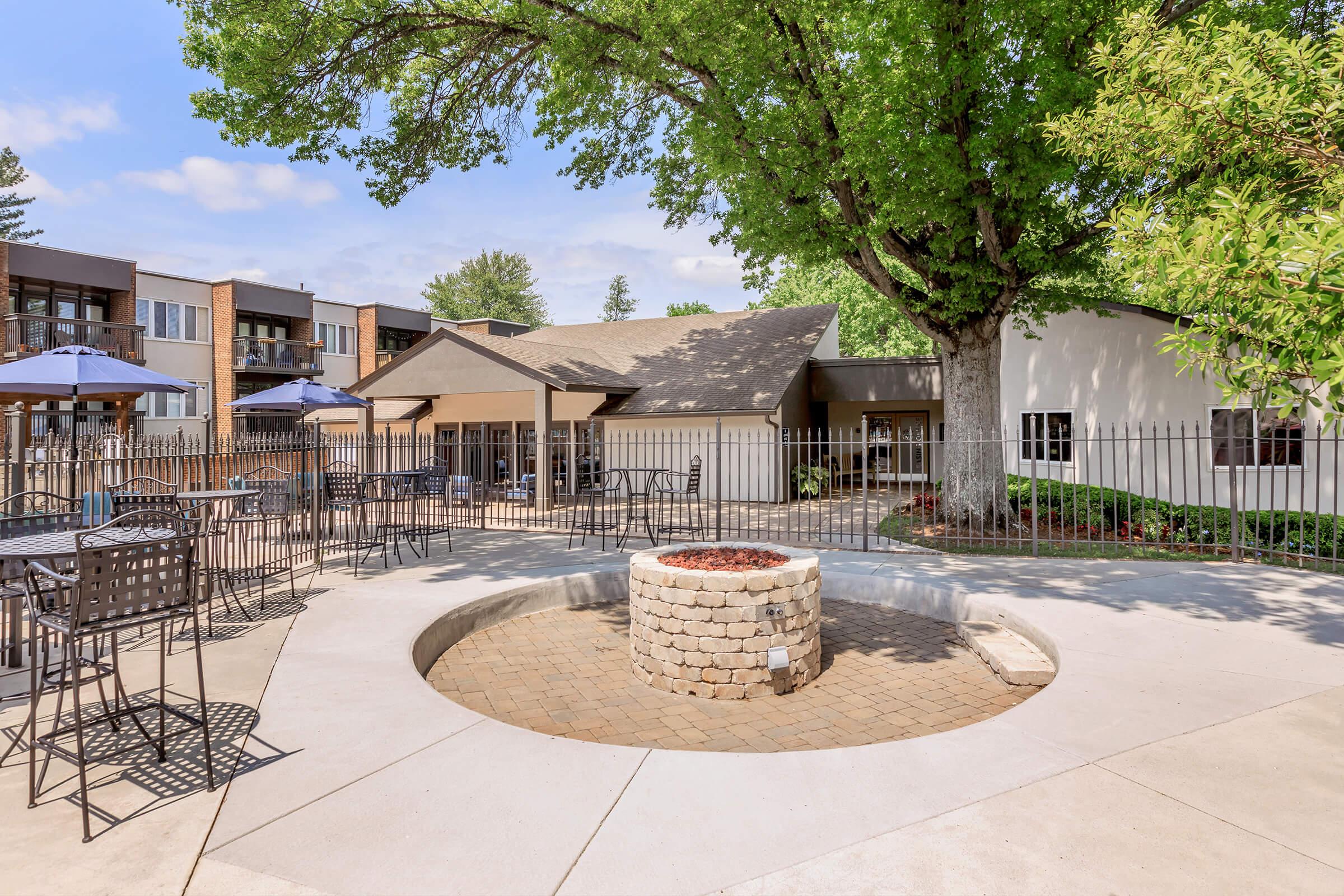 A landscaped outdoor area featuring a circular stone fire pit surrounded by paved seating. In the background, there are several tables with umbrellas and a modern building. Lush green trees provide shade, contributing to a relaxed, inviting atmosphere.