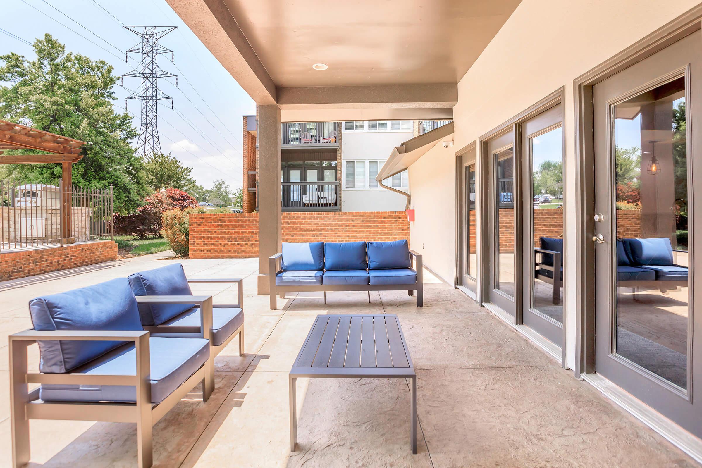 A modern outdoor seating area featuring blue cushioned furniture, including two chairs and a sofa, arranged around a small metal table. The space is located near a building, with a visible pathway and greenery in the background. Power lines are seen in the distance.