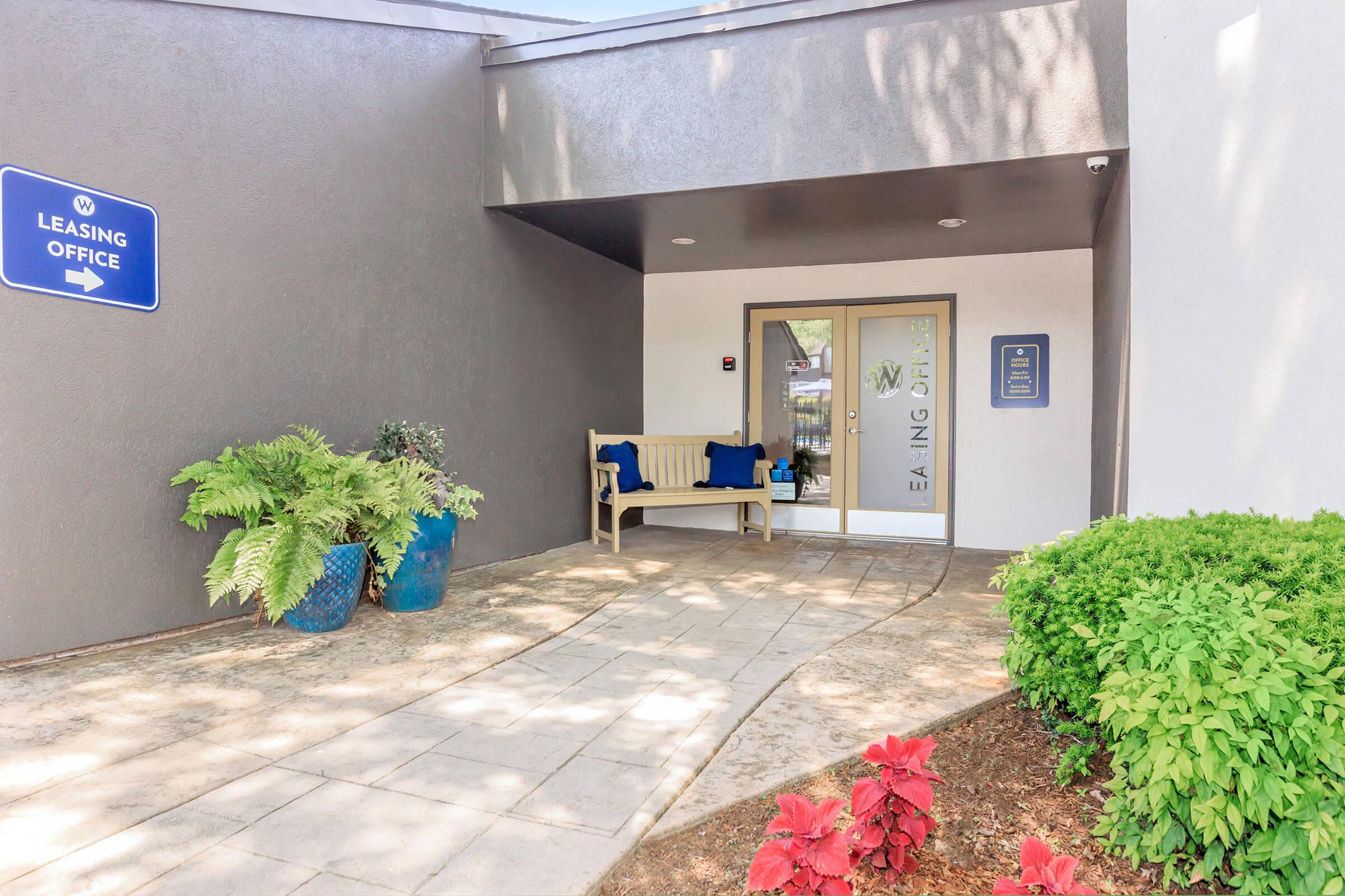 Entrance to a leasing office featuring a concrete pathway, a wooden bench, decorative blue planters with ferns, and vibrant red plants. The entrance is surrounded by a modern building facade with a sign indicating the leasing office.