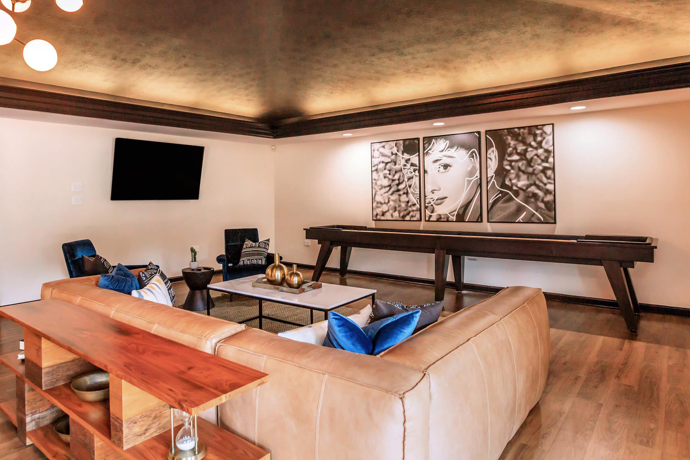 A spacious living room featuring a light brown leather sectional sofa with blue accent pillows, a large wooden coffee table, and a decorative centerpiece. There’s a flat-screen TV mounted on the wall, and an artwork of a person hanging above a long wooden console. Natural light fills the room, creating a warm atmosphere.