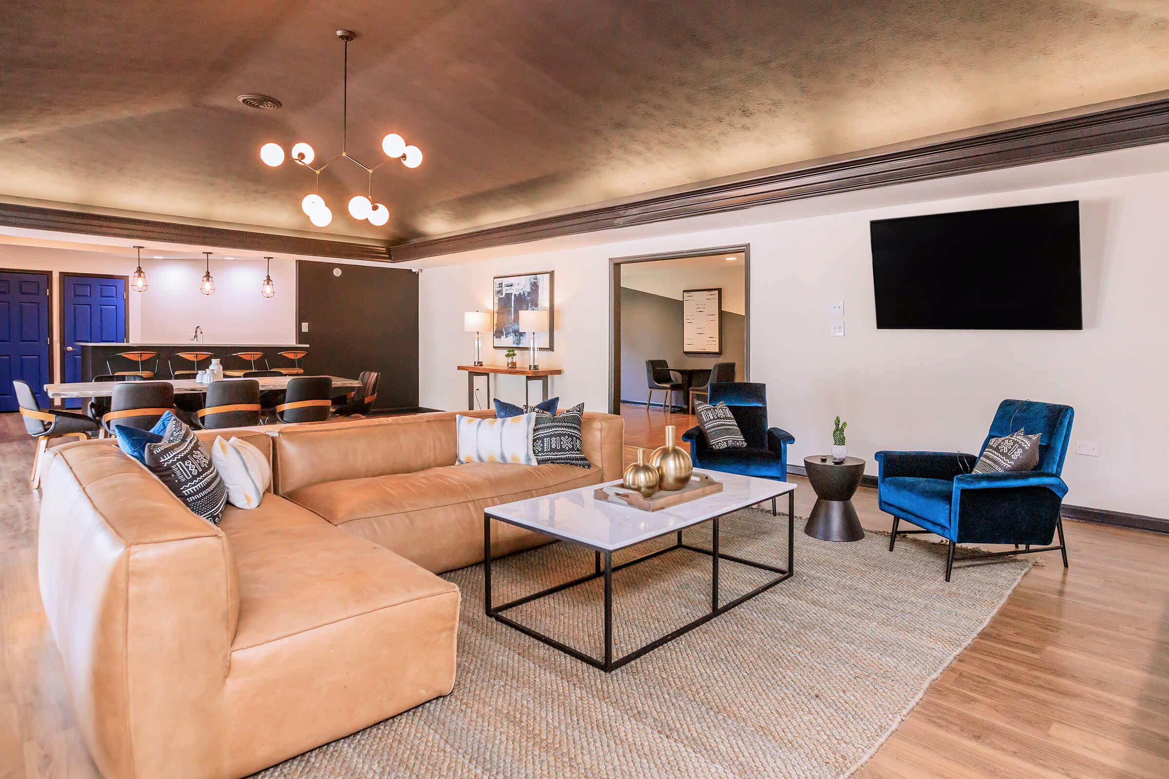 Modern living room featuring a large tan sectional sofa with decorative pillows, a sleek coffee table, and two blue accent chairs. Natural light floods the space, highlighting the stylish decor and contemporary design. A dark ceiling adds depth, and wall-mounted TV complements the elegant furnishings.