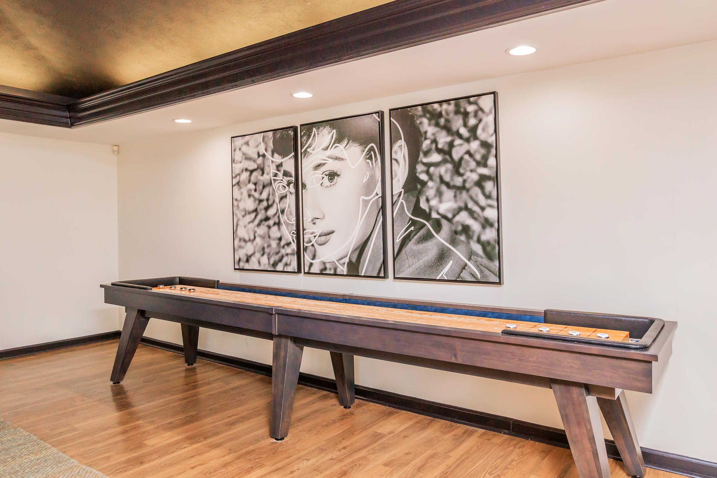 A game room featuring a wooden shuffleboard table in the foreground and two large black-and-white portraits on the wall above. The portraits depict stylized faces with intricate details. The room has wooden flooring and modern lighting, creating a vibrant yet relaxed atmosphere.