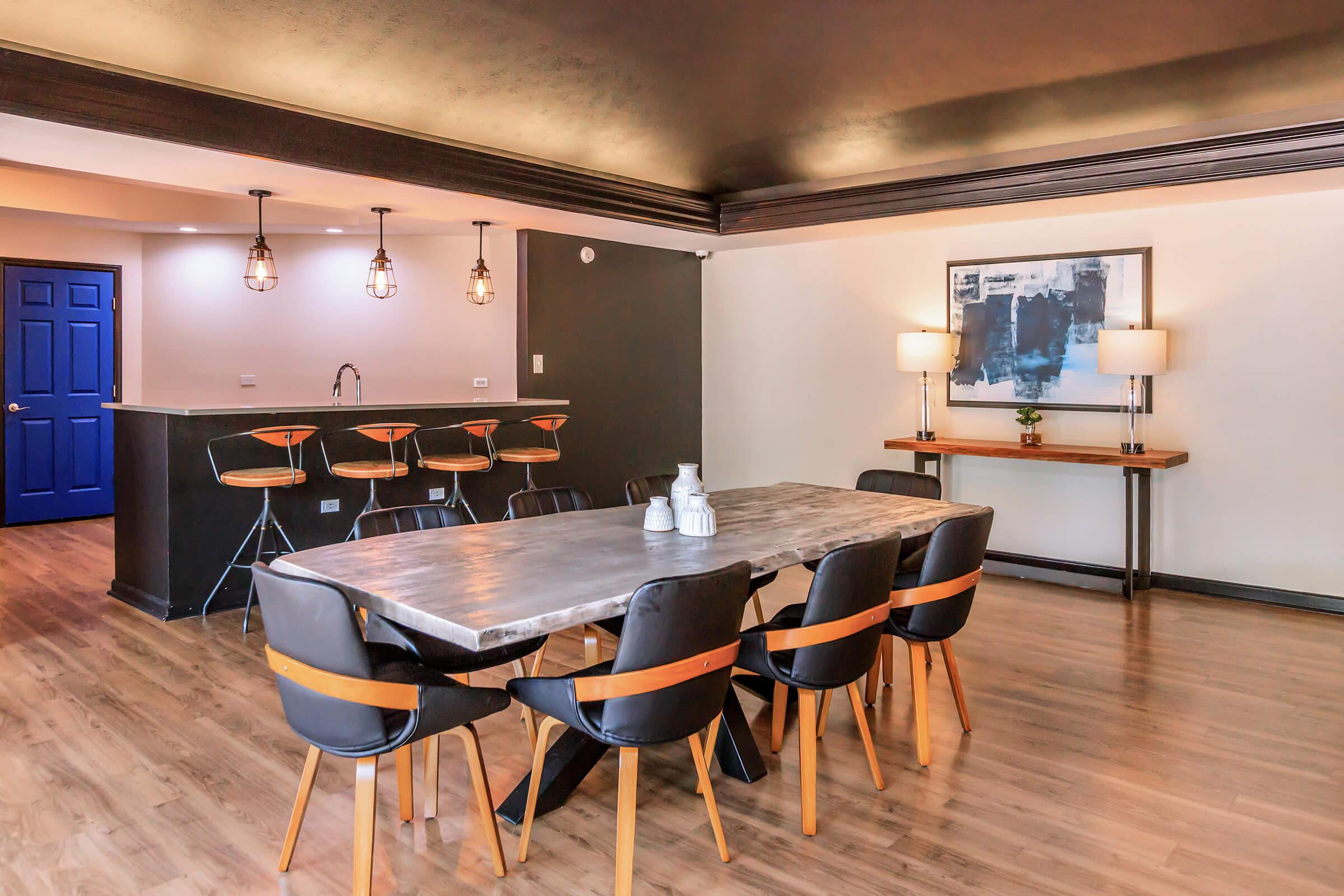 A modern dining area featuring a large wooden table surrounded by black and brown chairs. In the background, there is a dark kitchen bar with high stools, pendant lighting, and a blue door. The walls are painted in neutral tones, complemented by a large artwork and stylish table lamps.