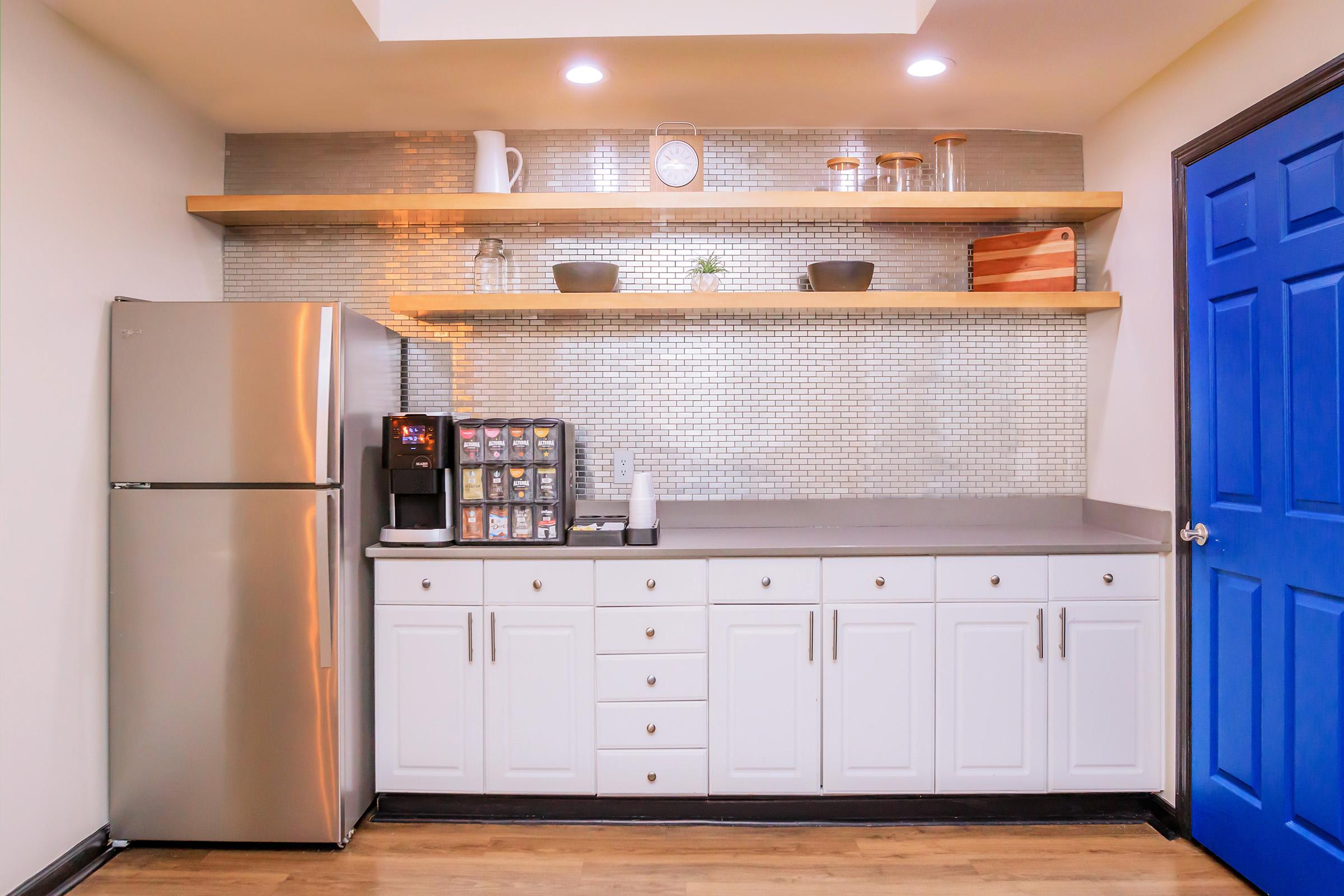 Modern kitchen with a stainless steel refrigerator, a coffee machine with a selection of coffee pods, and white cabinetry. Open wooden shelves display decorative items against a silver tiled backsplash. A bright blue door adds a pop of color to the warm wood flooring.