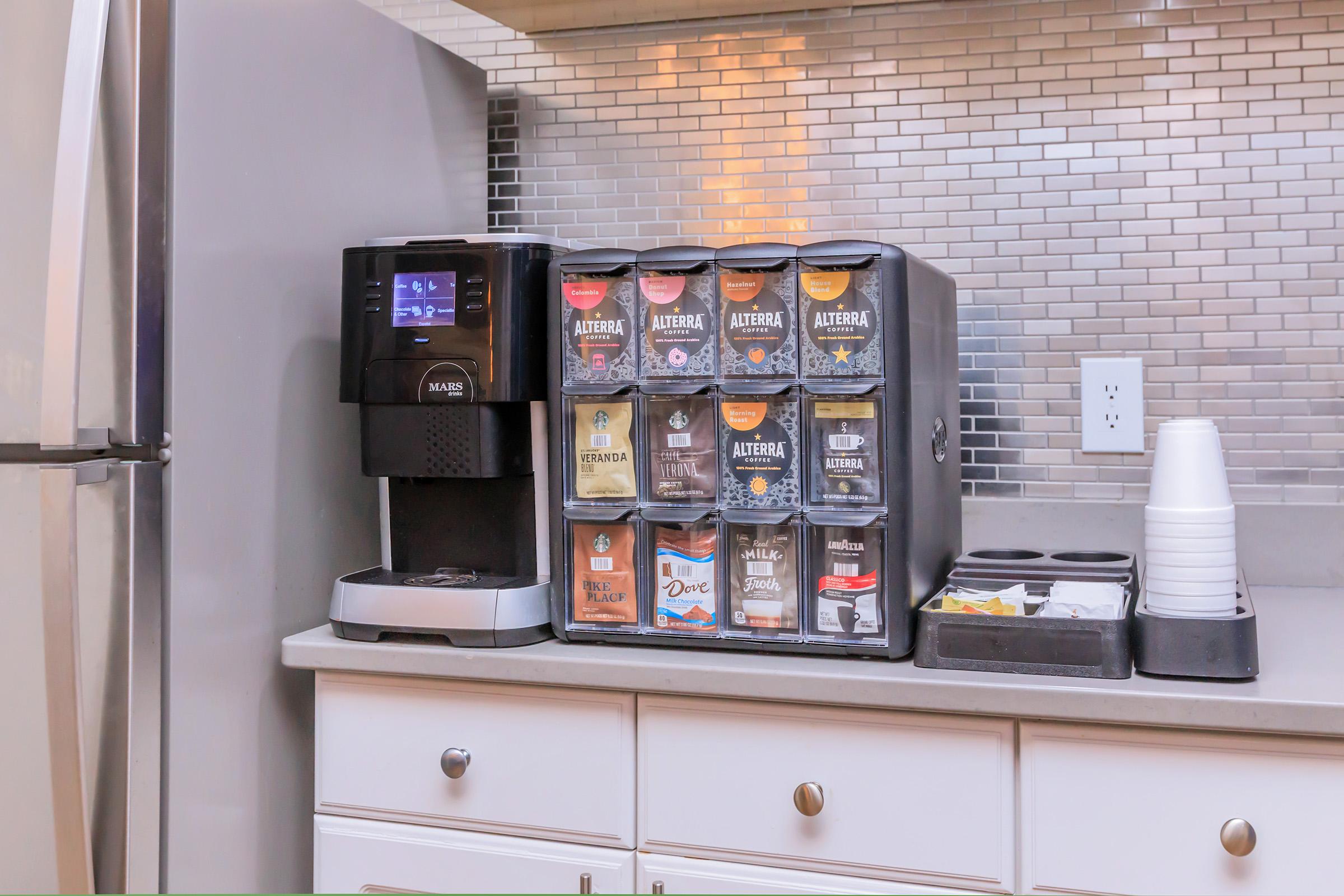 A modern kitchen counter featuring a sleek coffee machine next to a multi-compartment beverage dispenser. The dispenser holds various types of coffee and tea, with neatly organized cups and supplies nearby. The backsplash is made of reflective tiles, enhancing the contemporary design.