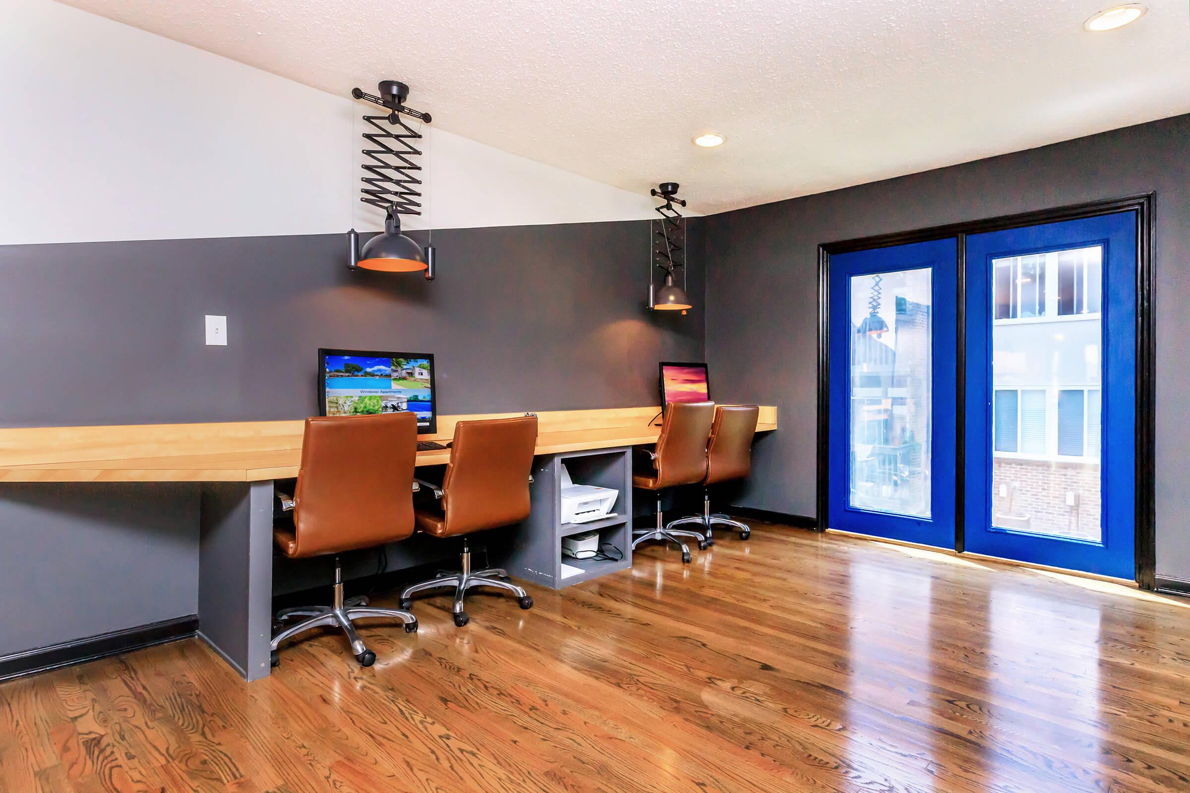 A modern workspace featuring a long wooden desk with three brown leather office chairs and two computers. The walls are painted in a two-tone gray design, and there are stylish pendant lights hanging above. Large blue-framed windows provide natural light and a view outside.