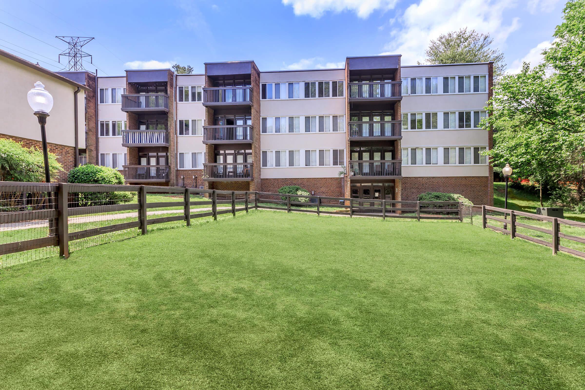 A well-maintained apartment building with multiple balconies, surrounded by a green lawn and a wooden fence. There are lamp posts along the path, and trees in the background under a clear blue sky.