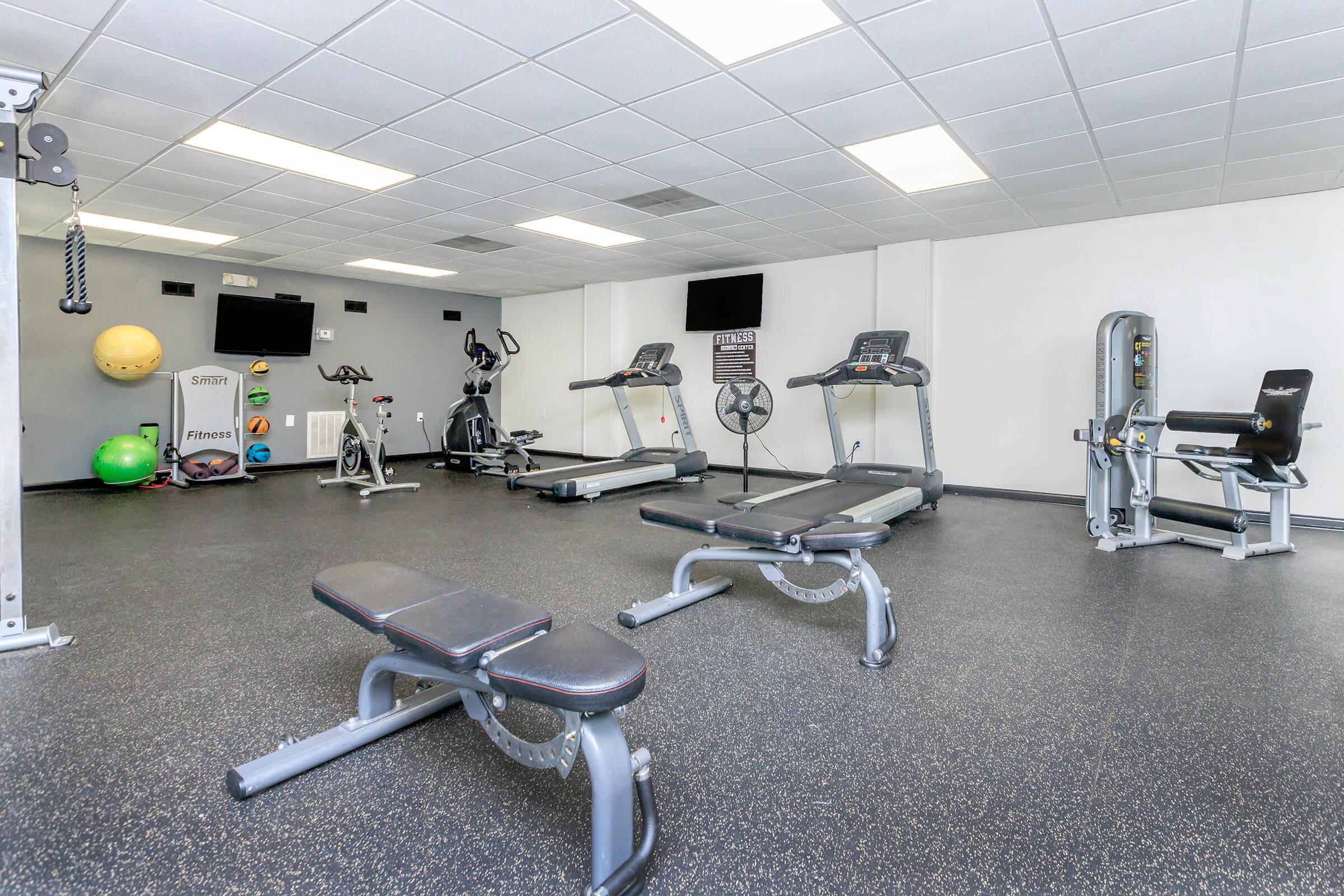 A clean and well-lit gym interior featuring various exercise equipment, including treadmills, a stationary bike, weight benches, and a multi-gym machine. Colorful exercise balls are visible, along with a fan and multiple screens displaying fitness-related information. The flooring is dark rubber for safety and comfort.