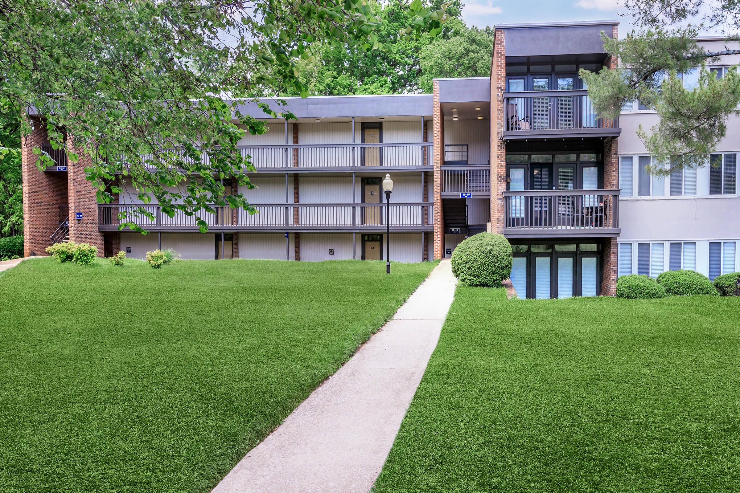 A modern apartment building with multiple balconies, surrounded by lush green grass and neatly trimmed bushes. A paved walkway leads to the entrance, and trees provide shade in the background, creating a pleasant residential area.
