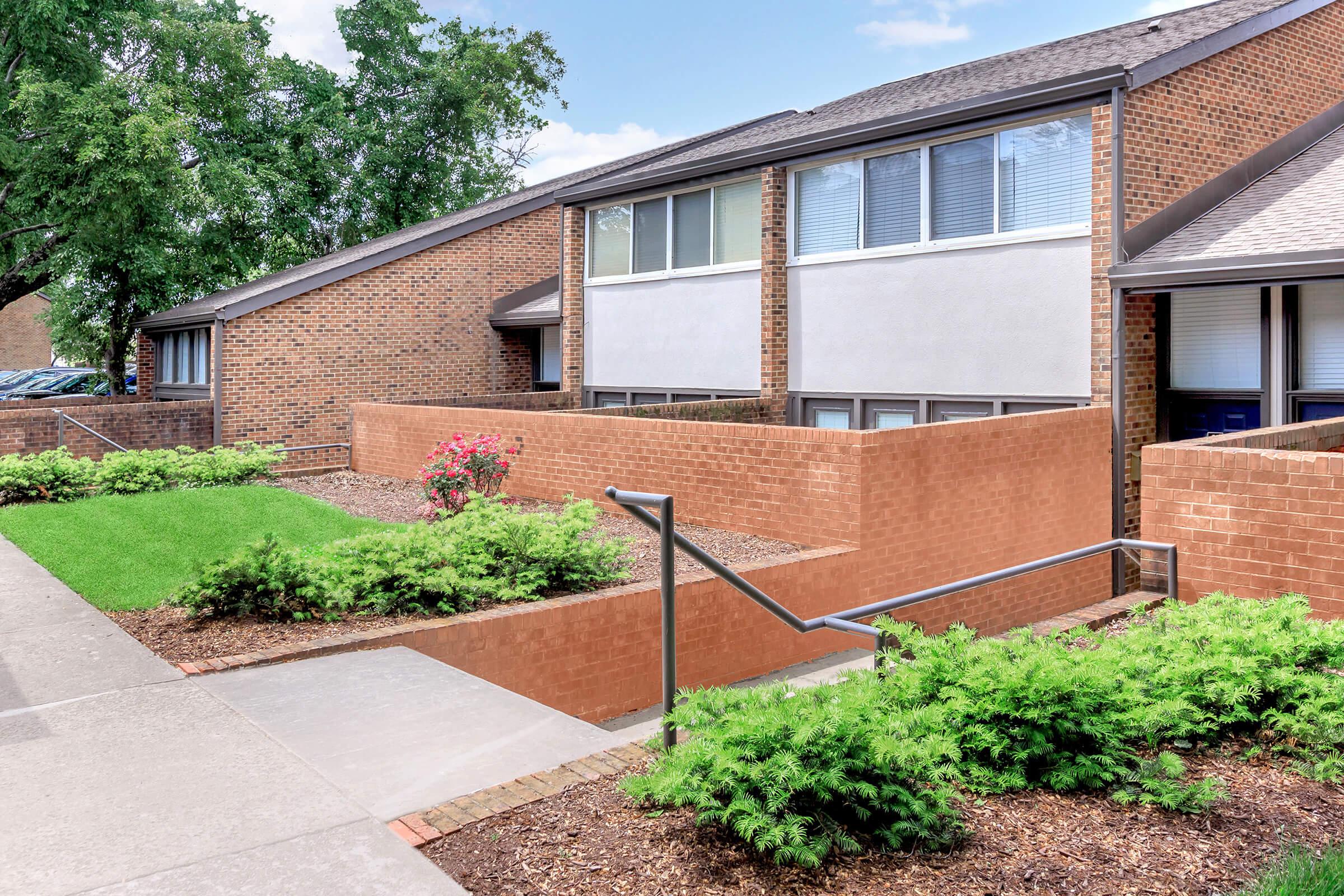 A well-maintained exterior of a residential building featuring brick walls, landscaped greenery, and a concrete pathway. The setting includes flowers and shrubs, along with a ramp for accessibility, creating a pleasant and inviting atmosphere.