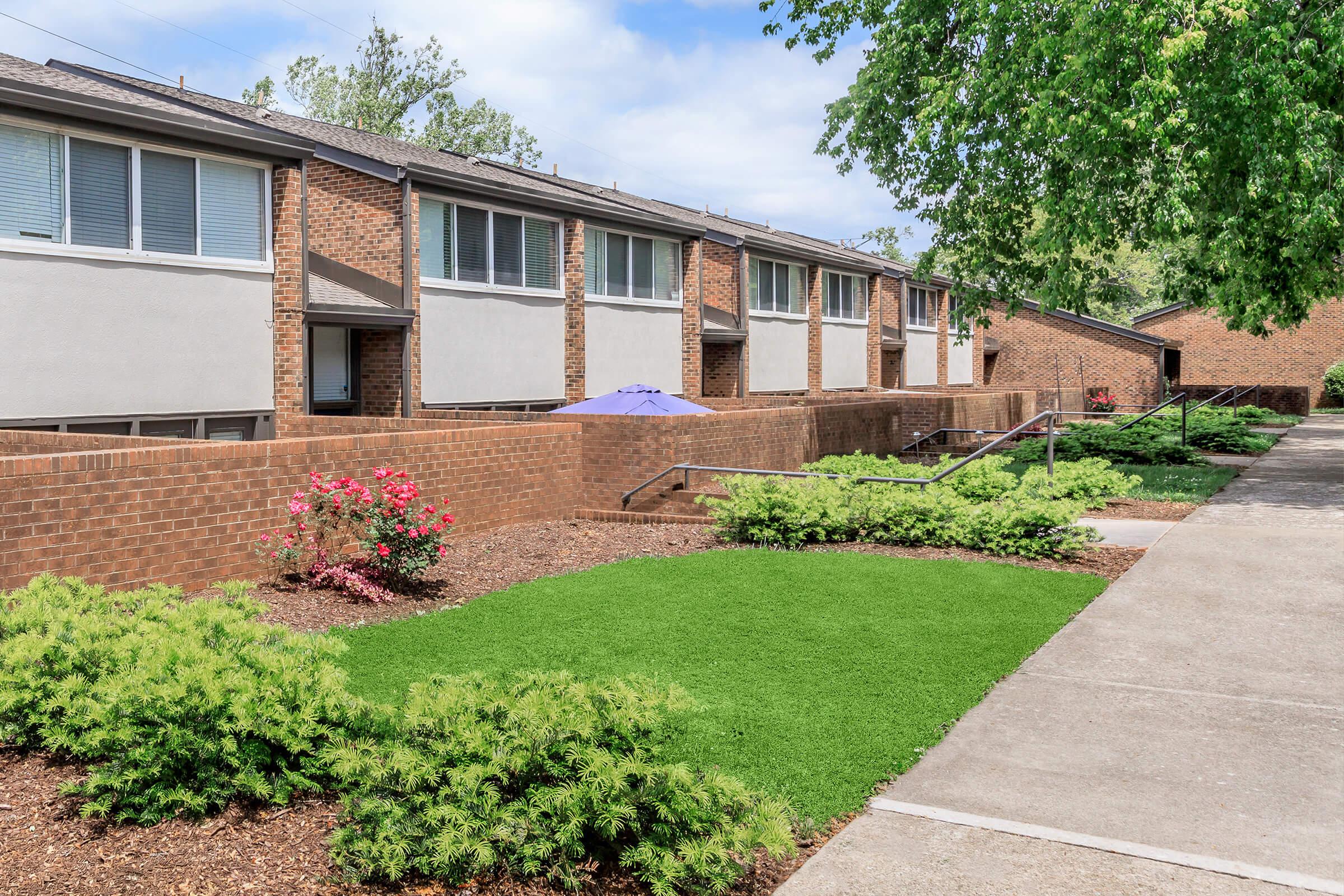 A view of a residential area featuring two-story brick apartment buildings. In the foreground, there is a walkway bordered by neatly manicured grass and shrubs. A purple umbrella sits in a small outdoor area among flowering plants. The scene is bright and well-maintained, with a clear sky above.