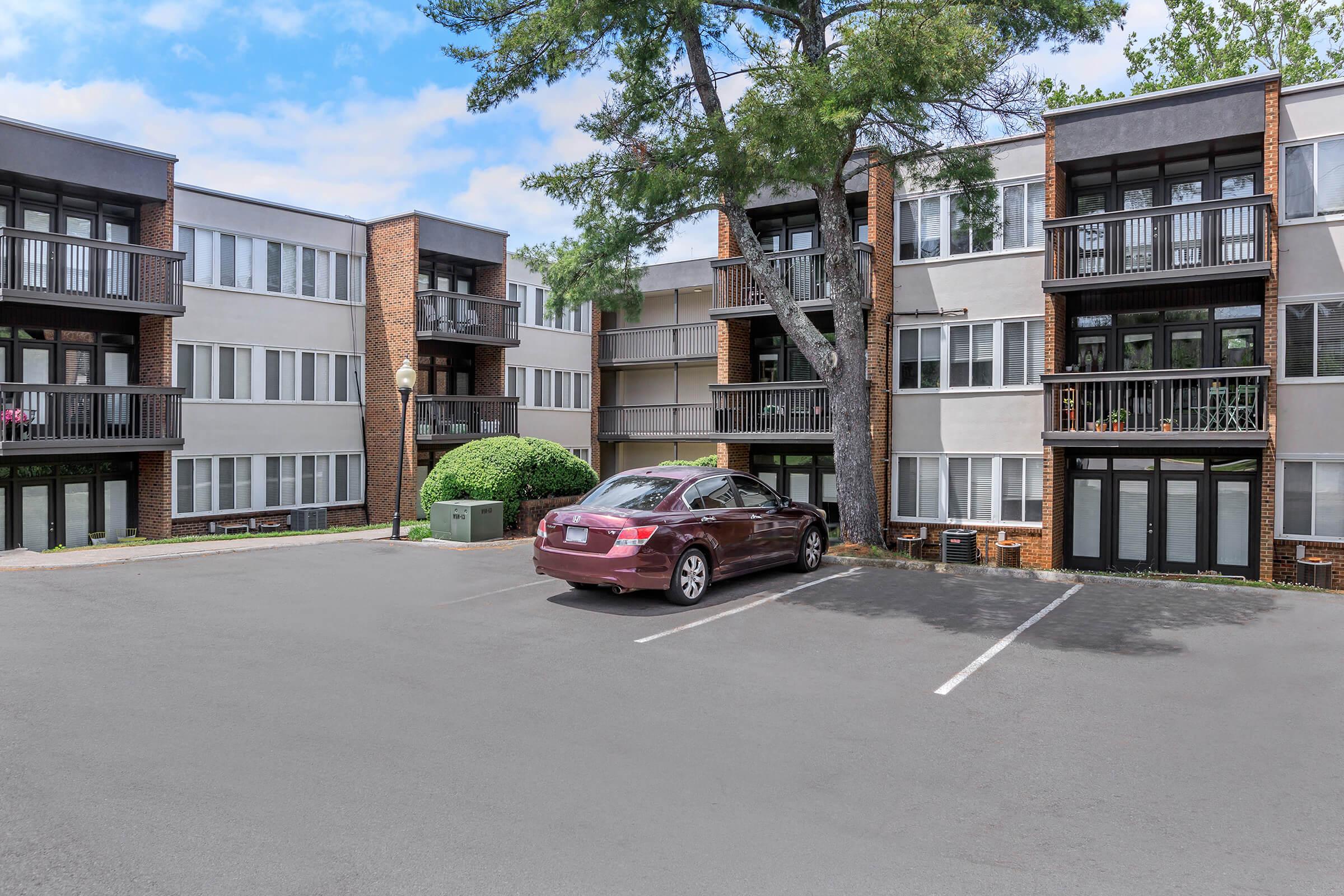 A view of a residential apartment complex with two buildings, featuring balconies and multiple windows. In the foreground, a parking lot with a maroon sedan parked. Lush greenery and trees are visible around the buildings against a clear blue sky.