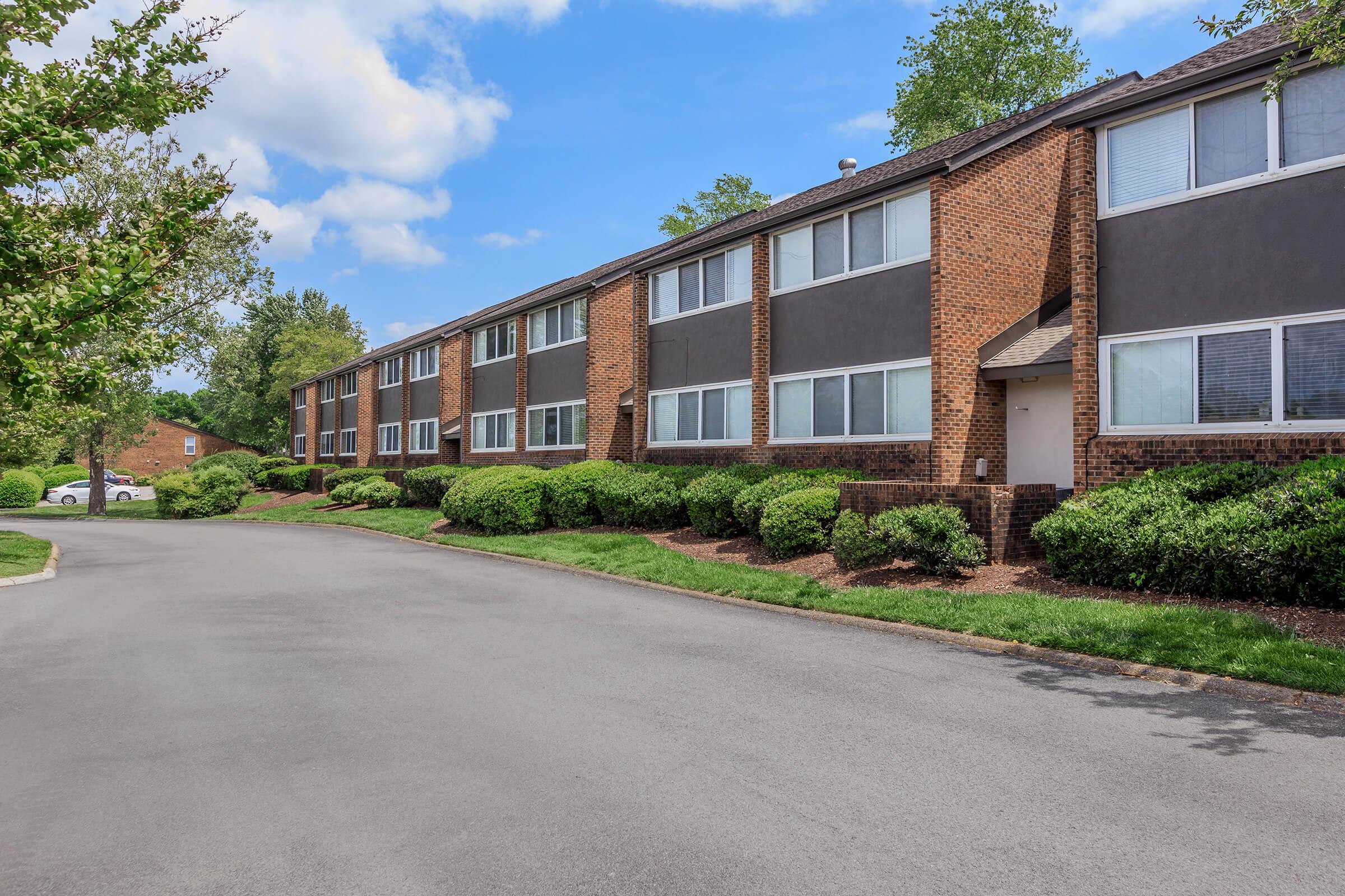 A row of brown brick apartment buildings with green shrubs and trees lining a paved driveway. The sky is partly cloudy, creating a bright atmosphere. The scene suggests a well-maintained residential area.