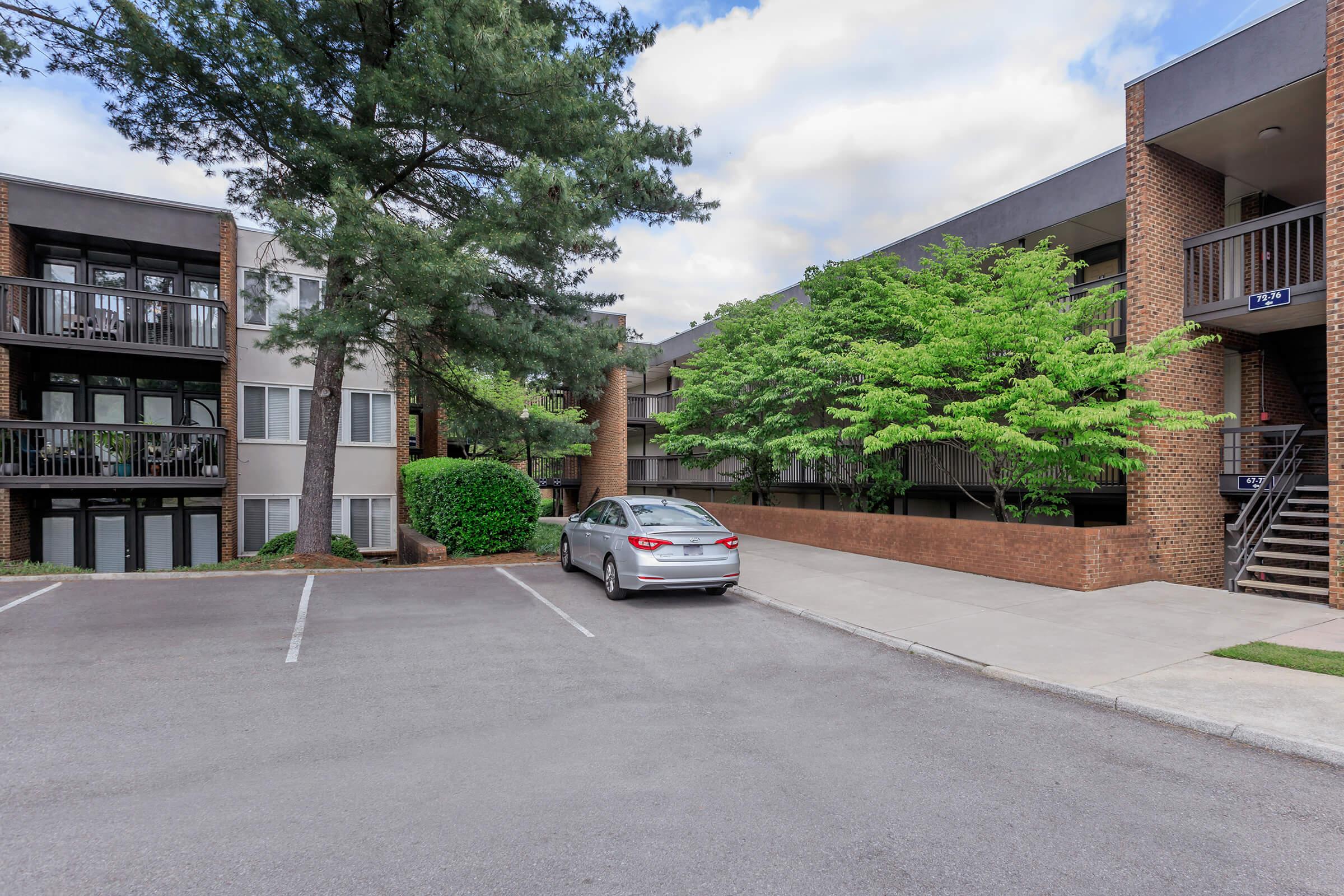 A residential apartment complex featuring multiple buildings, greenery, and a parking area. A silver car is parked in the foreground on a paved lot, with trees and shrubs visible nearby. The buildings have balconies and are set against a cloudy sky.