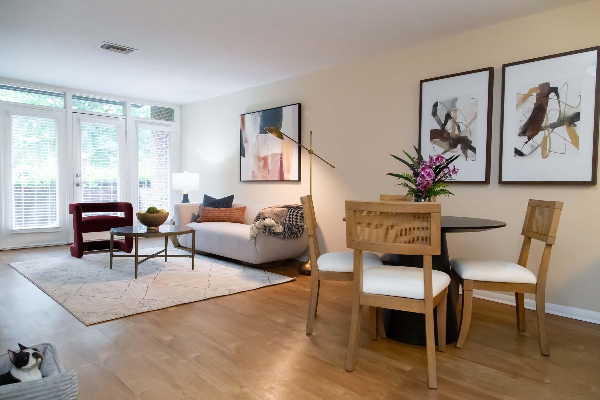 A cozy living room featuring a light-colored sofa with decorative pillows, a round dining table with chairs, a plant centerpiece, and two framed abstract art pieces on the wall. A large window lets in natural light, and a cat is resting on the floor. Warm wooden flooring adds to the inviting atmosphere.
