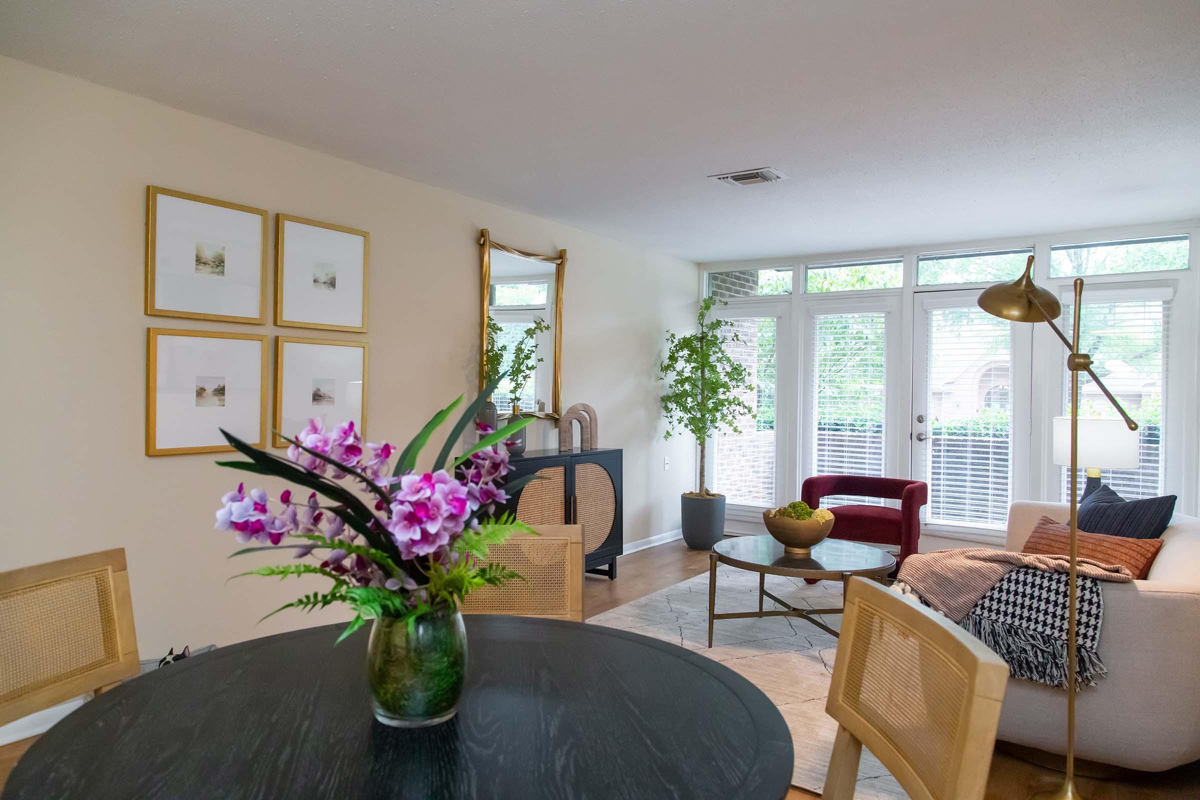 A cozy living space featuring a round black dining table with a floral centerpiece, a beige sofa adorned with throw pillows, and a red armchair. Natural light floods in through large windows, highlighting indoor plants and framed artwork on the walls, creating a warm and inviting atmosphere.
