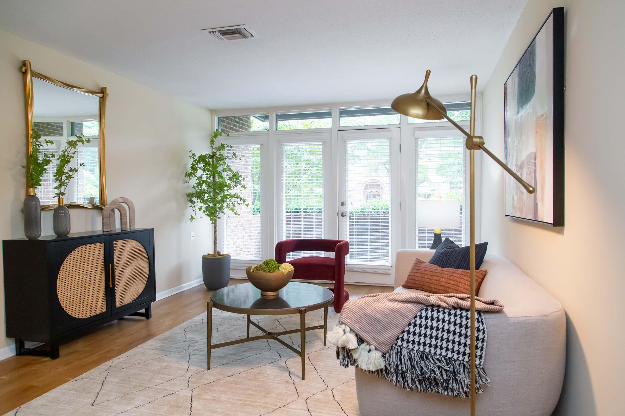 A modern living room featuring a light-colored sofa adorned with a patterned throw blanket, a round glass coffee table with a decorative bowl, and a stylish black cabinet. A large mirror reflects natural light from the windows, and potted plants add a touch of greenery to the space.