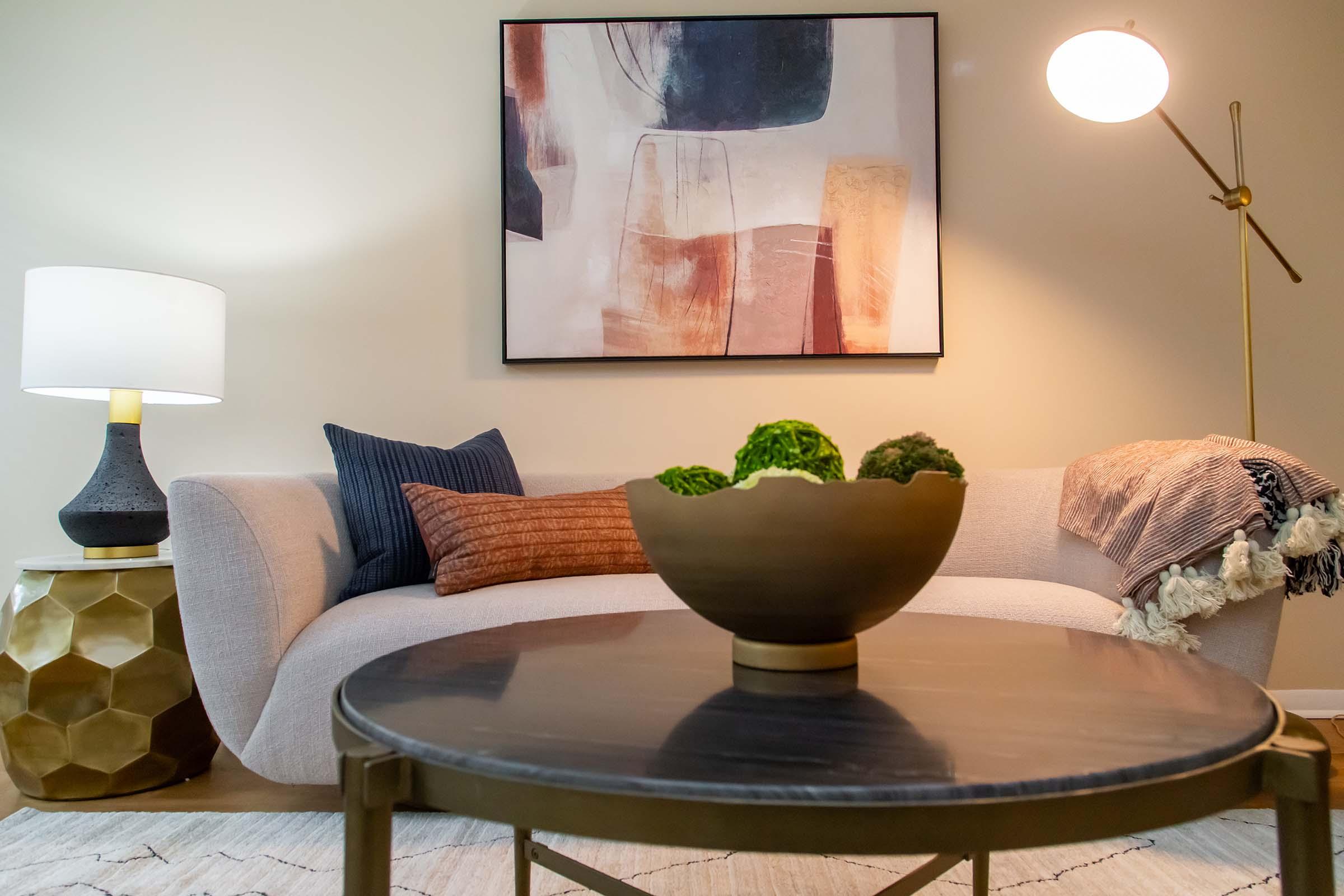 A cozy living room setup featuring a light gray sofa with decorative pillows, a round coffee table with a decorative bowl, and a stylish gold lamp. The wall is adorned with an abstract painting in muted colors, and a throw blanket is draped over the sofa.