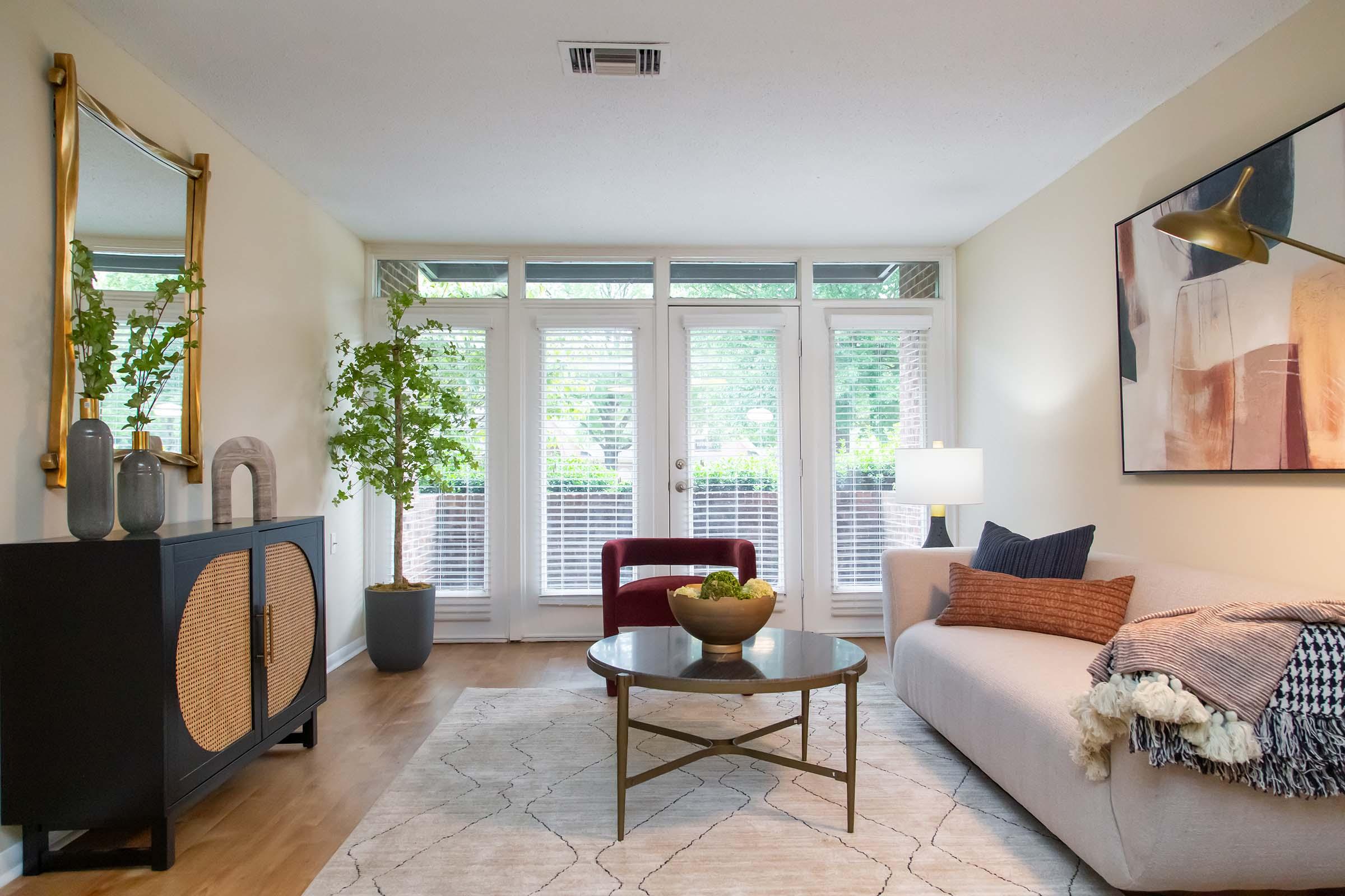 A cozy living room featuring a light-colored sofa with decorative pillows, a round coffee table with a green centerpiece, a modern entertainment unit, and large windows providing natural light. Potted plants add a touch of greenery, and contemporary artwork hangs on the wall, creating an inviting atmosphere.