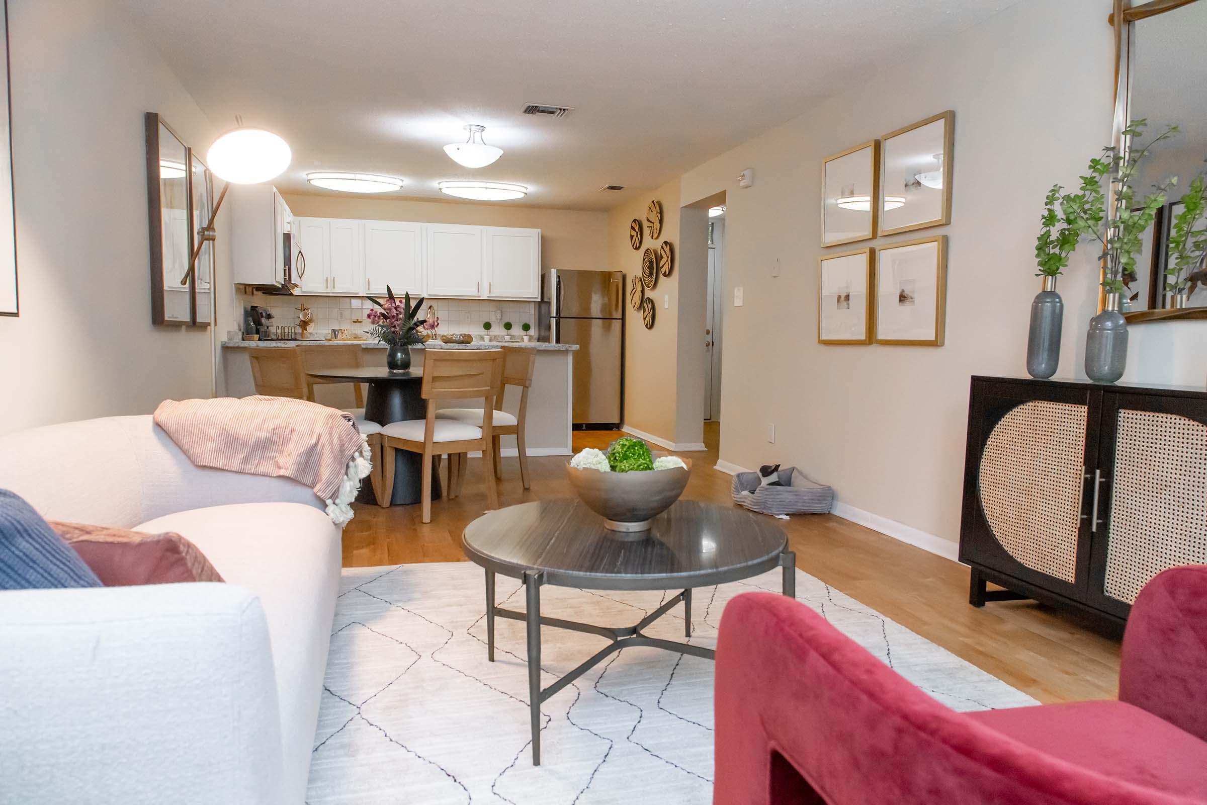 A cozy living room featuring a light-colored sofa with pillows, a round coffee table with a decorative bowl, and two red chairs. In the background, there's a modern kitchen with white cabinets and stainless steel appliances, along with decorative wall art and small plants adding a touch of elegance.