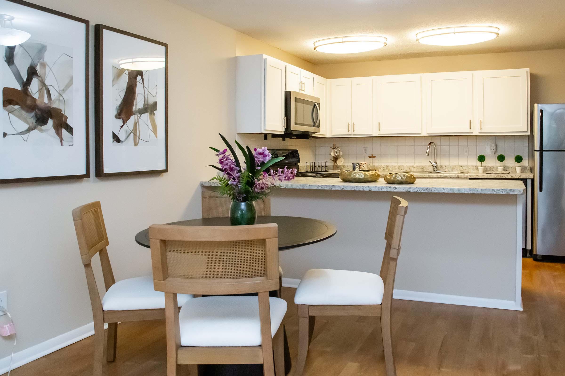 A modern kitchen and dining area featuring a round black table with four wooden chairs, a vase of purple flowers on the table, and light-colored cabinetry in the background. The kitchen includes a microwave, stove, and stainless-steel refrigerator, with decorative art on the walls. Bright, neutral colors create a welcoming atmosphere.