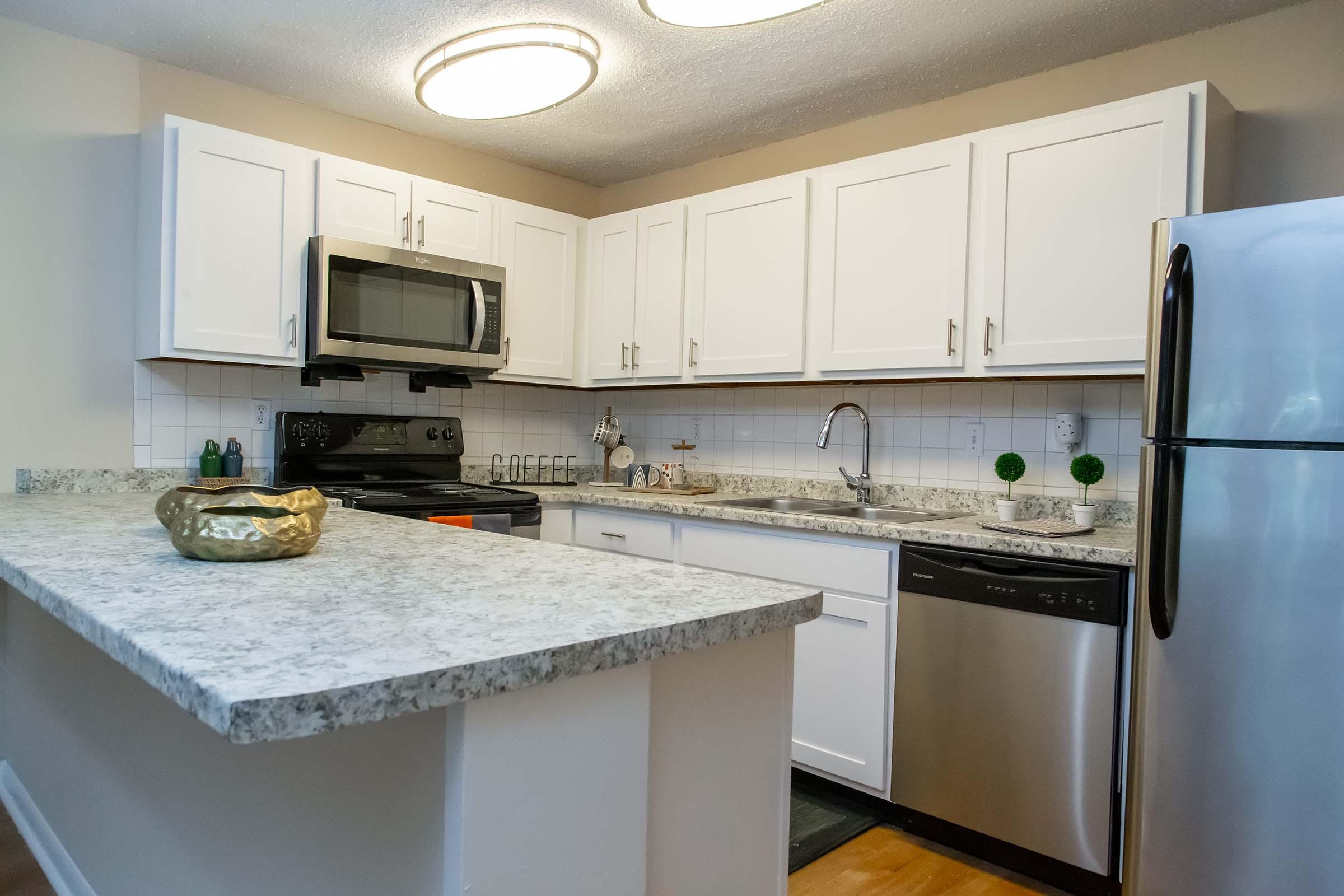 Modern kitchen featuring white cabinets, stainless steel appliances including a microwave, oven, and dishwasher. A granite countertop complements the kitchen island, which has a decorative bowl. The space is well-lit with overhead lighting and includes a sink and backsplash.