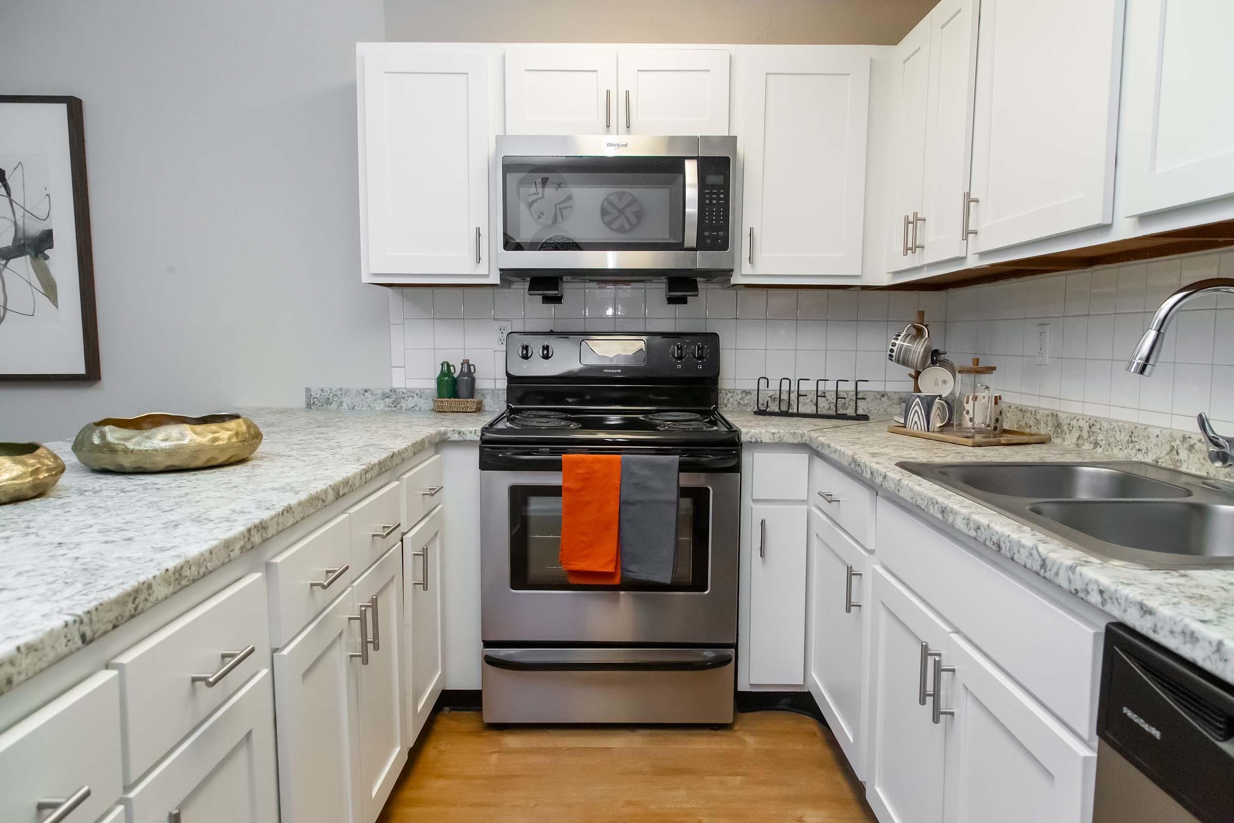 A modern kitchen featuring white cabinets, a gray granite countertop, a stainless steel oven, and a microwave above. There are decorative bowls on the counter, a sink, and kitchen utensils. Two towels hang from the oven in contrasting colors of orange and gray, creating a stylish and functional space.