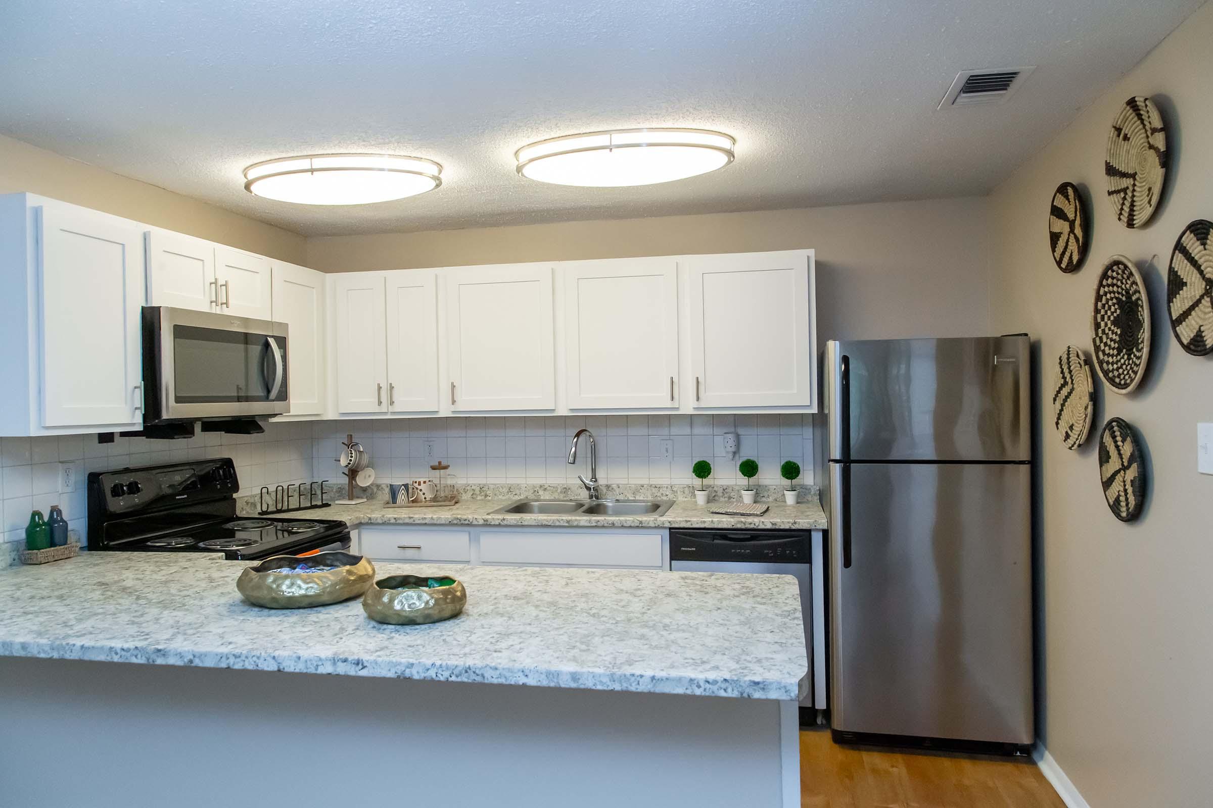 Modern kitchen featuring white cabinets, a stainless steel refrigerator, and a built-in microwave. The countertop is made of granite, adorned with two decorative bowls. Circular ceiling lights illuminate the space, while woven wall art adds a decorative touch.
