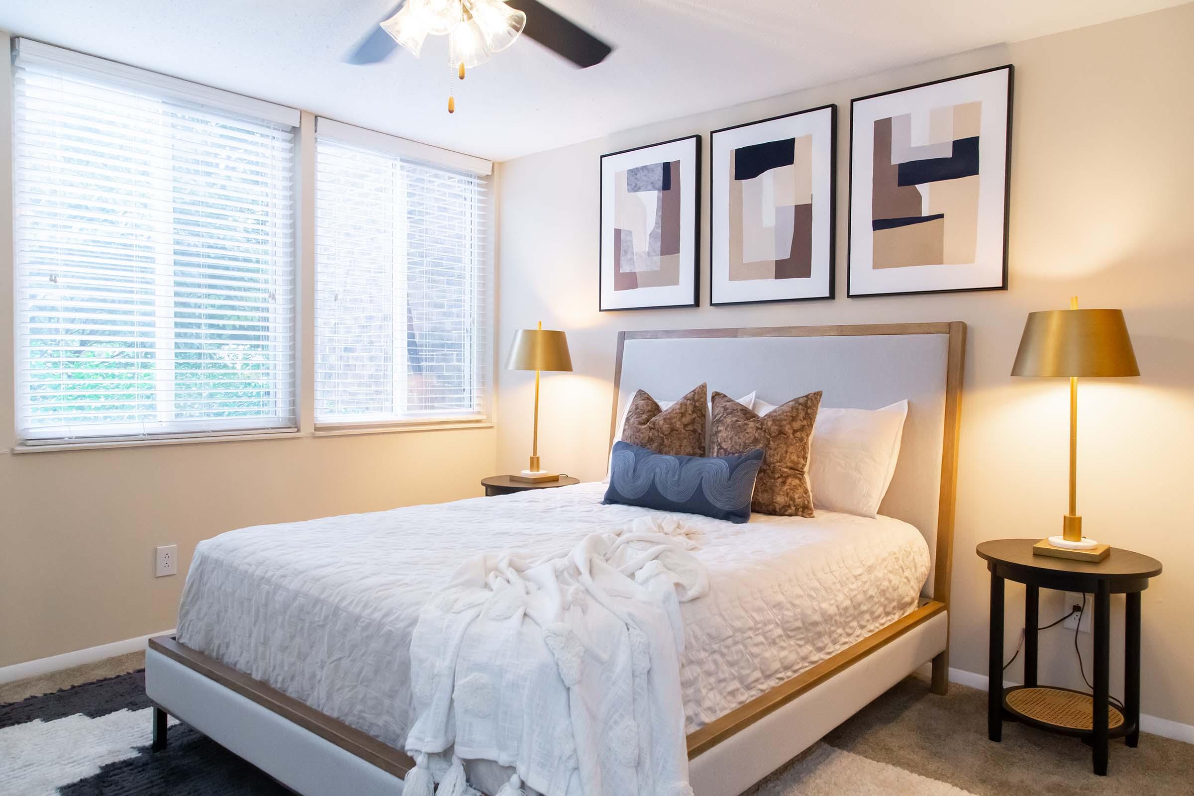 A cozy bedroom featuring a queen-sized bed with a light gray upholstered headboard, adorned with decorative pillows and a soft throw. The room has two bedside tables with stylish lamps, large windows with blinds, and three modern framed art pieces on the wall, creating a warm and inviting atmosphere.
