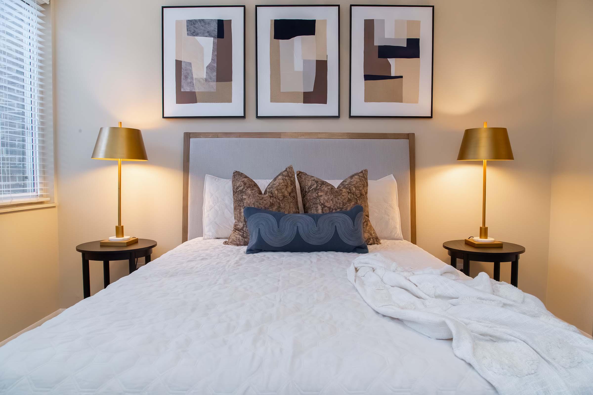 A cozy bedroom featuring a neatly made bed with decorative pillows, flanked by two stylish lamps on side tables. Above the bed, three abstract art pieces in neutral colors adorn the wall, complementing the warm ambiance of the room. Natural light streams in through a window with blinds.