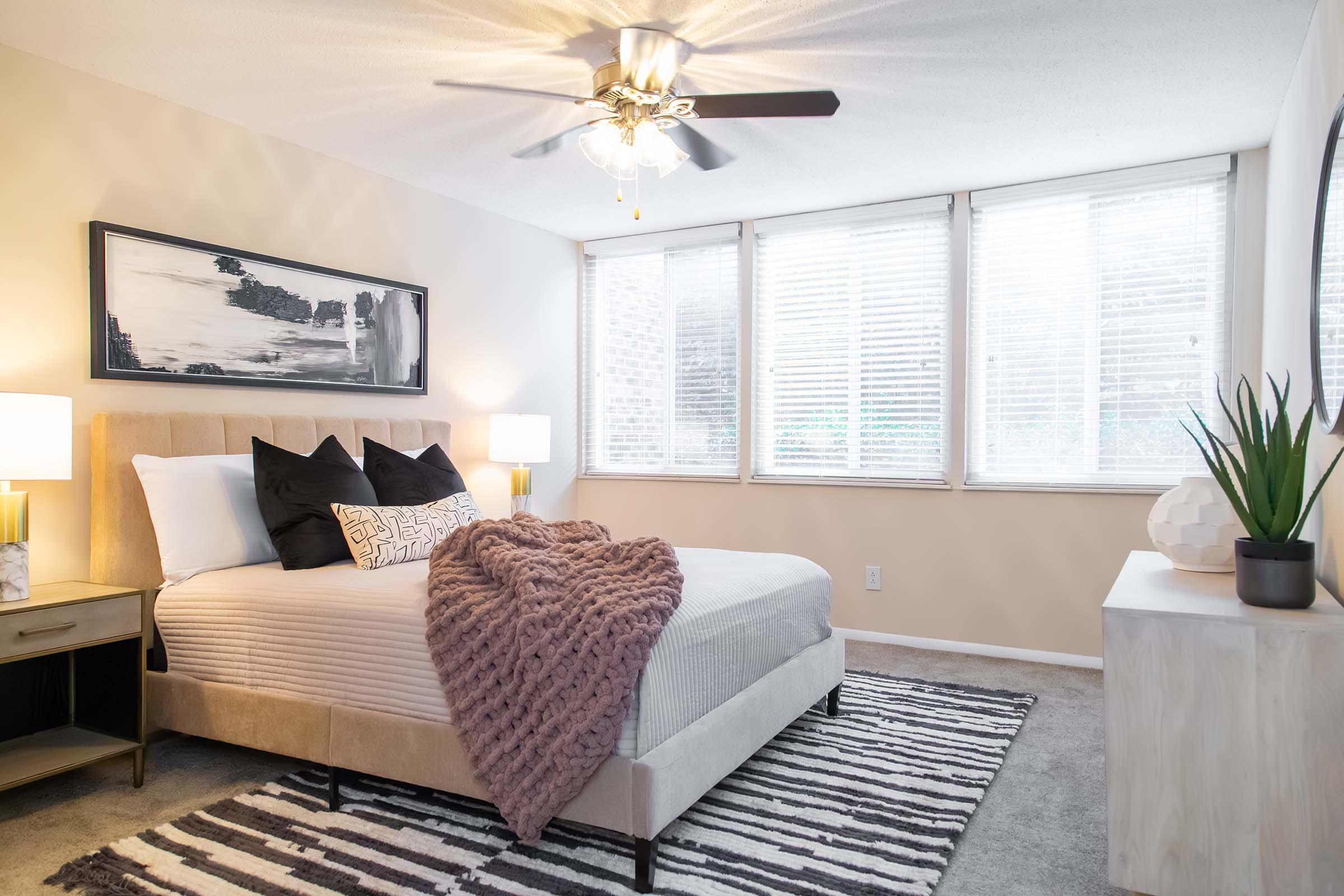 Cozy bedroom featuring a queen-sized bed with decorative pillows and a plush throw blanket. The room has large windows with blinds, a ceiling fan, two bedside lamps, and a stylish dresser. A modern black and white artwork hangs above the bed, and a potted plant adds a touch of greenery.