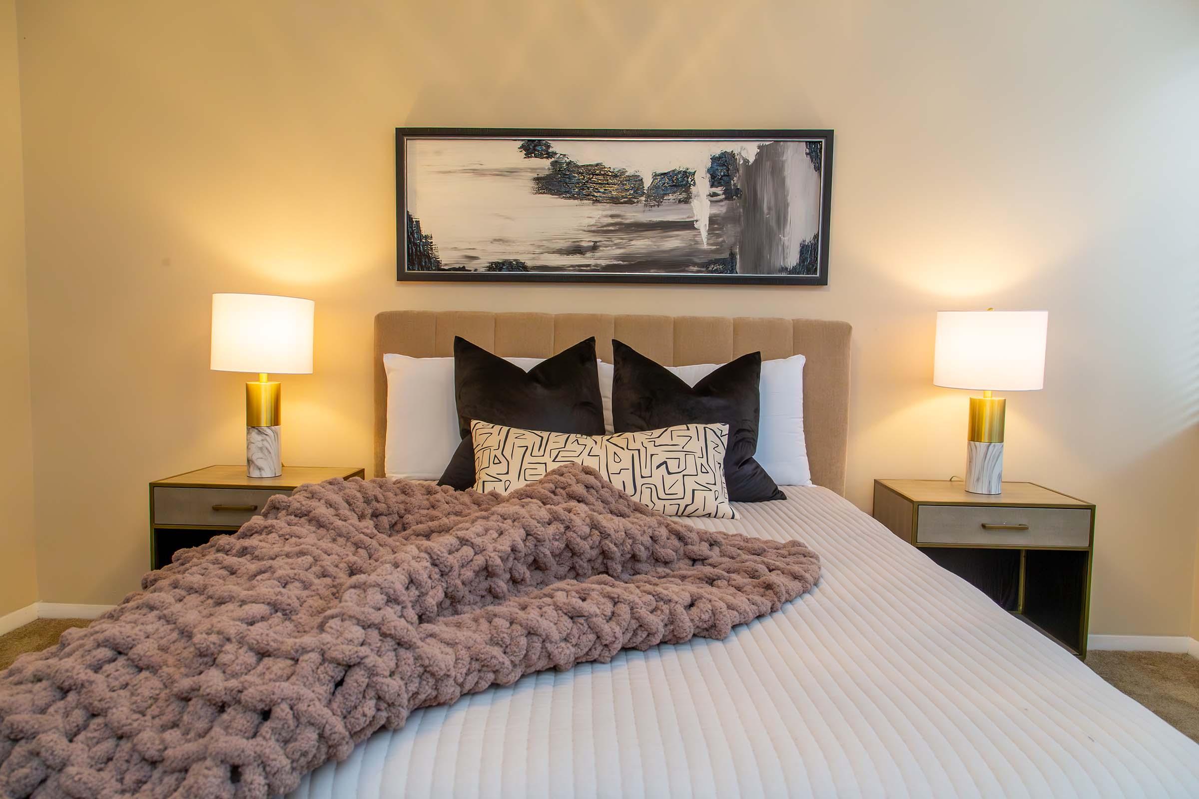 A cozy bedroom scene featuring a neatly made bed with a soft beige headboard. The bed is adorned with decorative pillows and a plush, knitted blanket. On either side of the bed, there are nightstands with lamps, and a large abstract painting hangs above the headboard, enhancing the warm ambiance.
