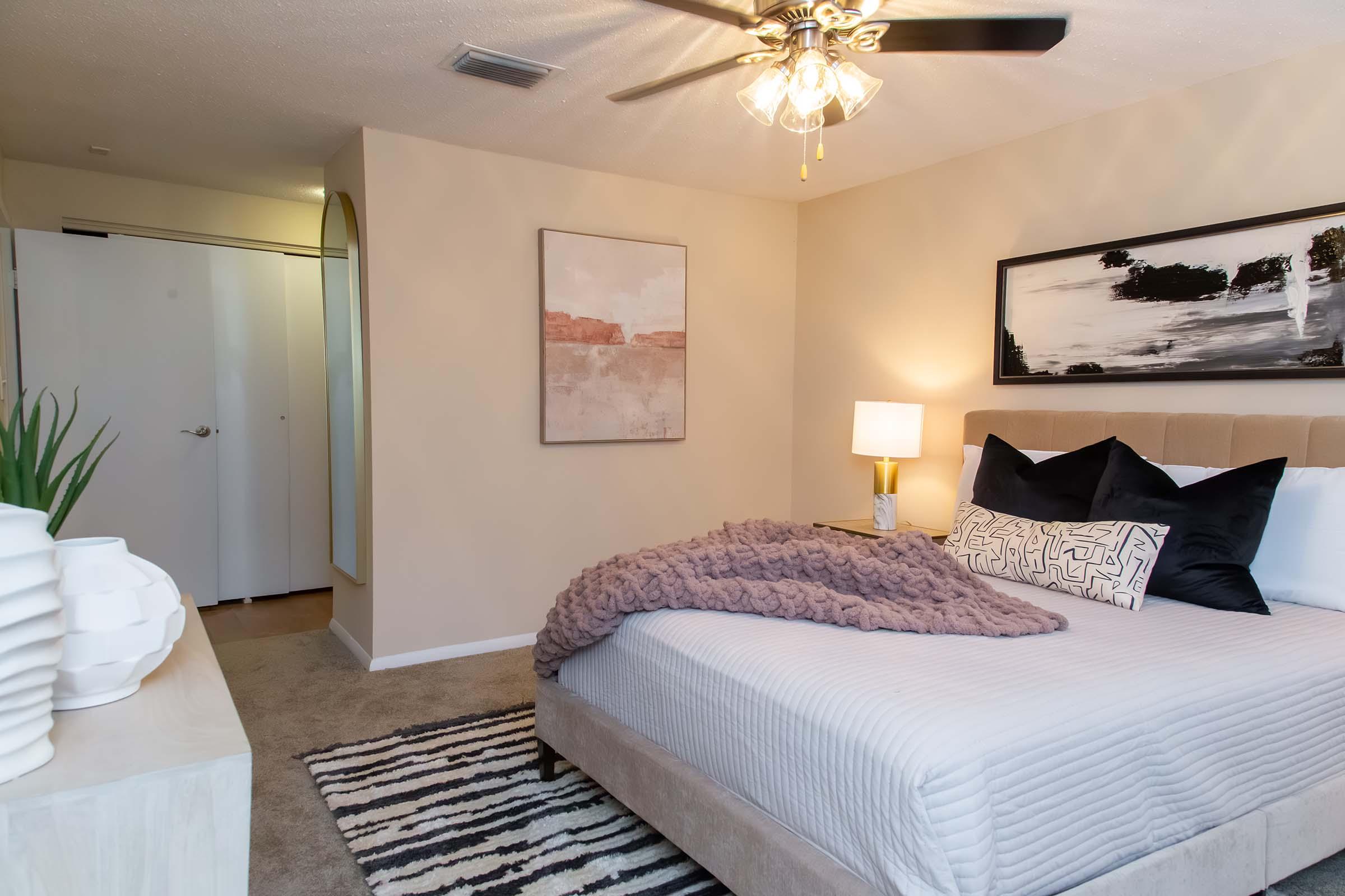 Cozy bedroom featuring a plush bed with a beige headboard and decorative pillows, a soft purple throw blanket, a nightstand with a lamp, and a wall art piece. The room has light beige walls, a ceiling fan, and a stylish area rug, creating a warm and inviting atmosphere. An open closet door is visible in the background.