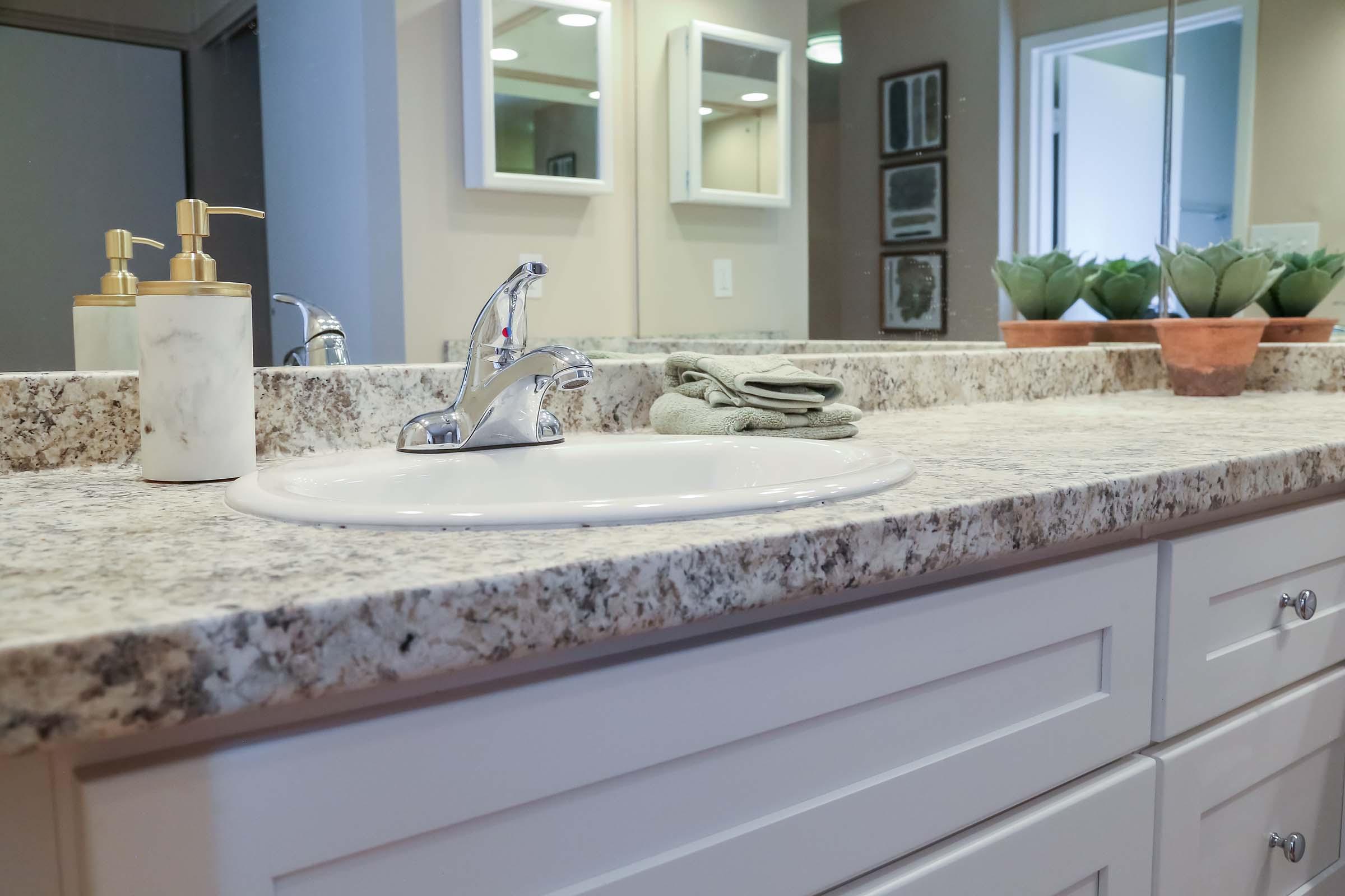 A bathroom countertop featuring a white sink with a shiny faucet, surrounded by decorative items including a marble soap dispenser, a folded towel, and small potted plants. Mirrors are positioned above the sink, reflecting the room's neutral colors and modern design.