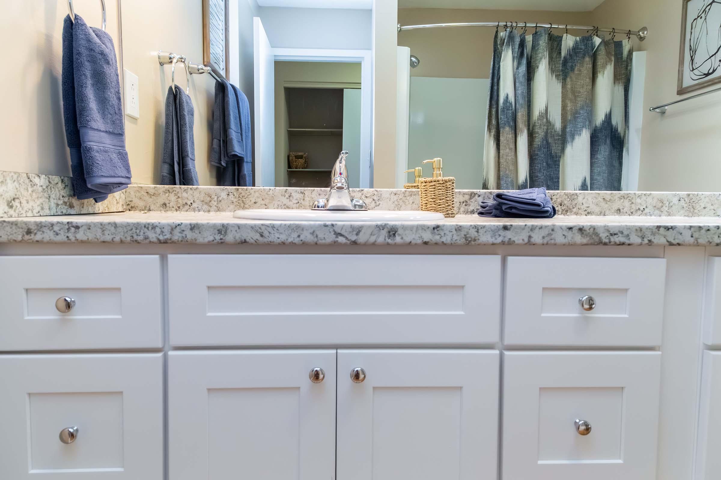 A bathroom countertop featuring a white sink with a polished chrome faucet, granite surface, and neatly arranged items. There are blue towels hanging on the wall and a decorative basket on the counter. The background includes a shower curtain with a geometric pattern and a partially visible cabinet.