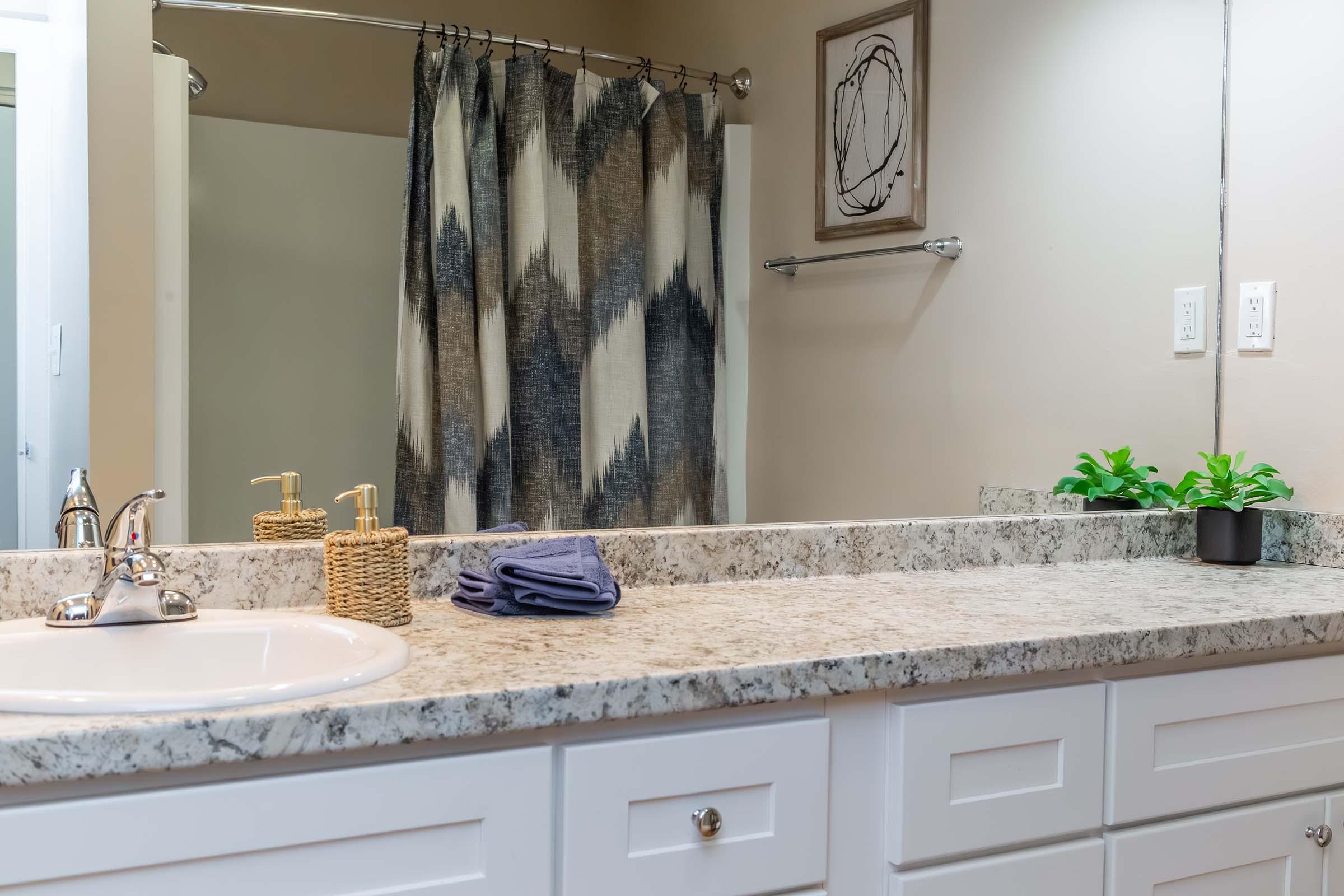 A modern bathroom countertop featuring a white sink with polished chrome faucet, two soap dispensers, a small potted plant, and neatly folded blue towels. The background shows a patterned shower curtain and a framed abstract art piece, complemented by neutral wall colors and a clean, minimalistic design.