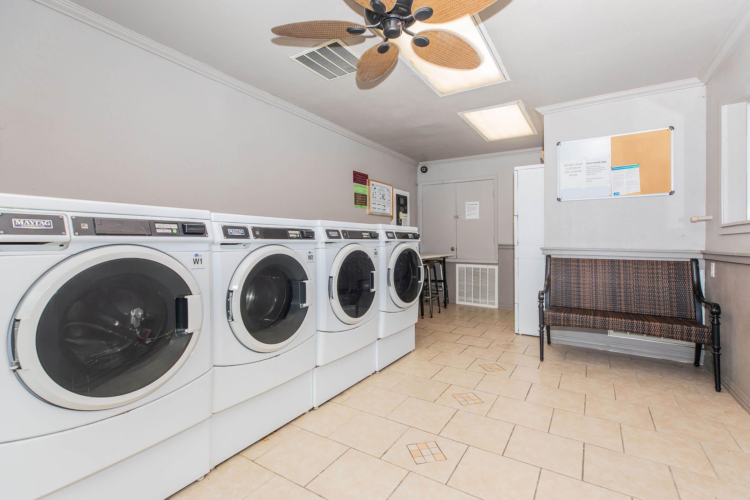 A clean laundry room featuring four front-loading washing machines, a ceiling fan, a bench, and informational bulletin boards on the walls. The tile floor adds to the spacious, well-lit environment.