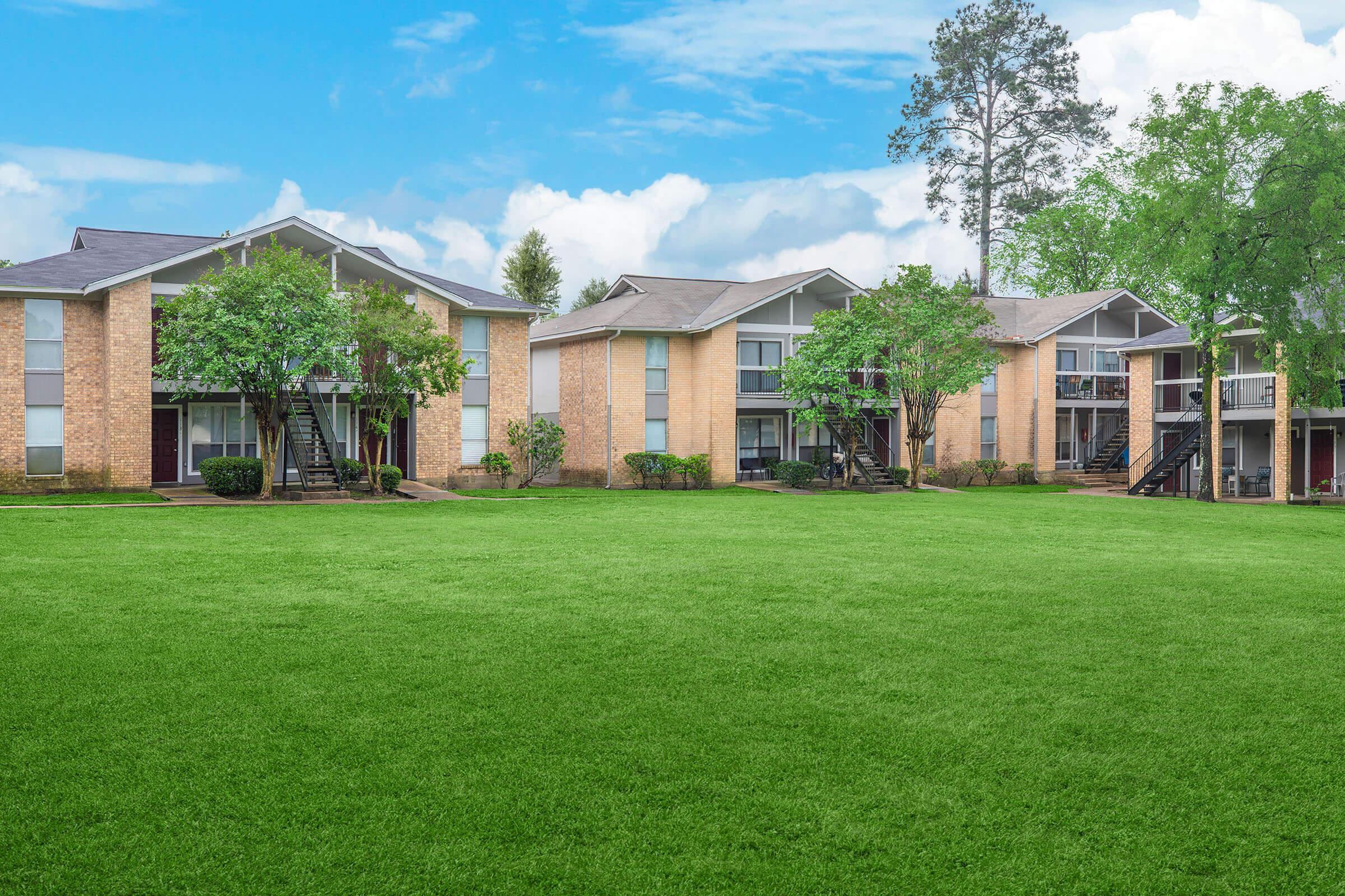 A landscaped residential area featuring multiple two-story apartment buildings with brick exteriors. Each building has small balconies and is surrounded by well-maintained green lawns and trees. The sky is blue with a few clouds, creating a bright and inviting atmosphere.