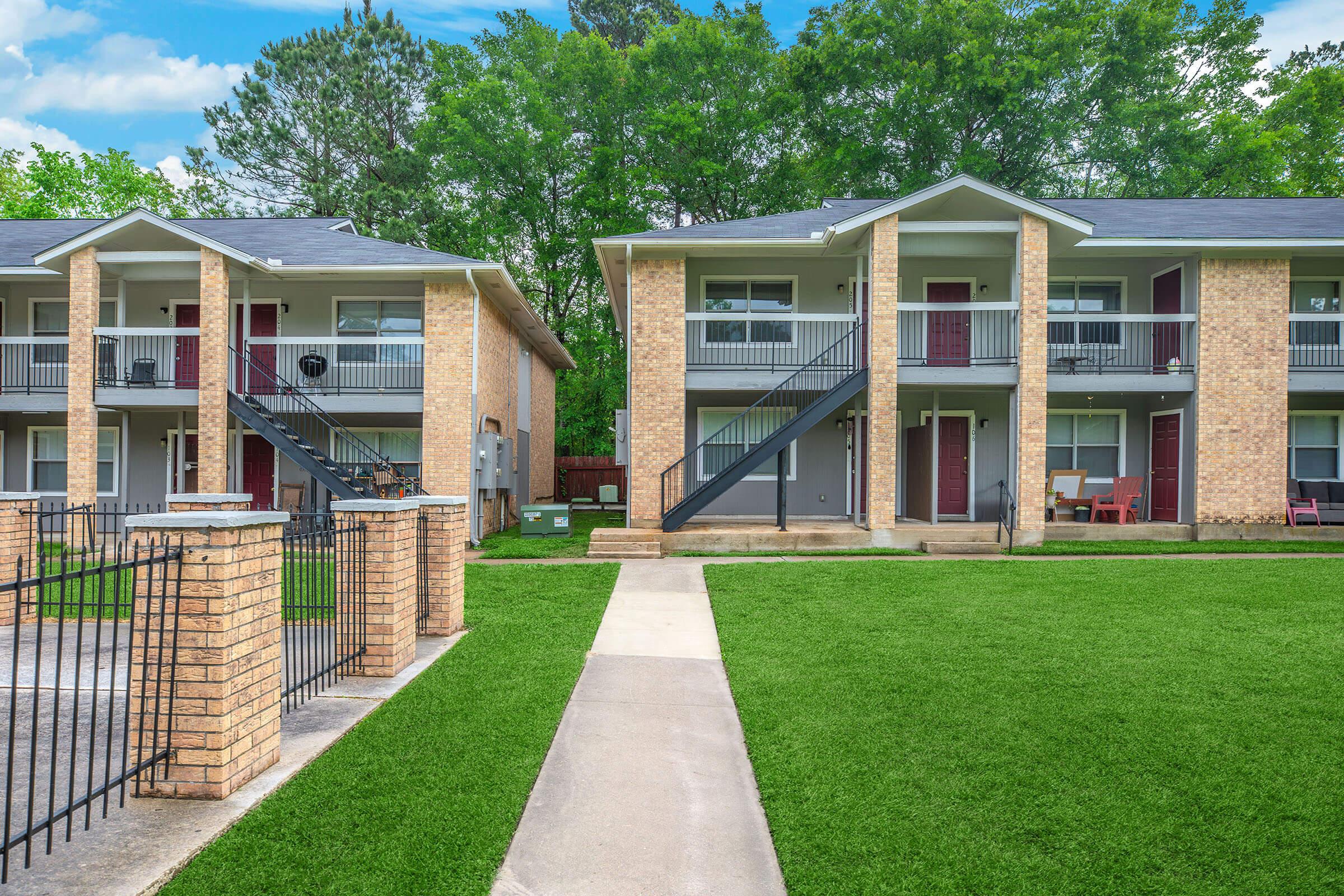 A view of two identical two-story apartment buildings with brick exteriors, separated by a manicured lawn and a walkway. Each building features a staircase leading to the upper level apartments, with balconies partially visible. Lush green trees are in the background, adding to the inviting atmosphere.