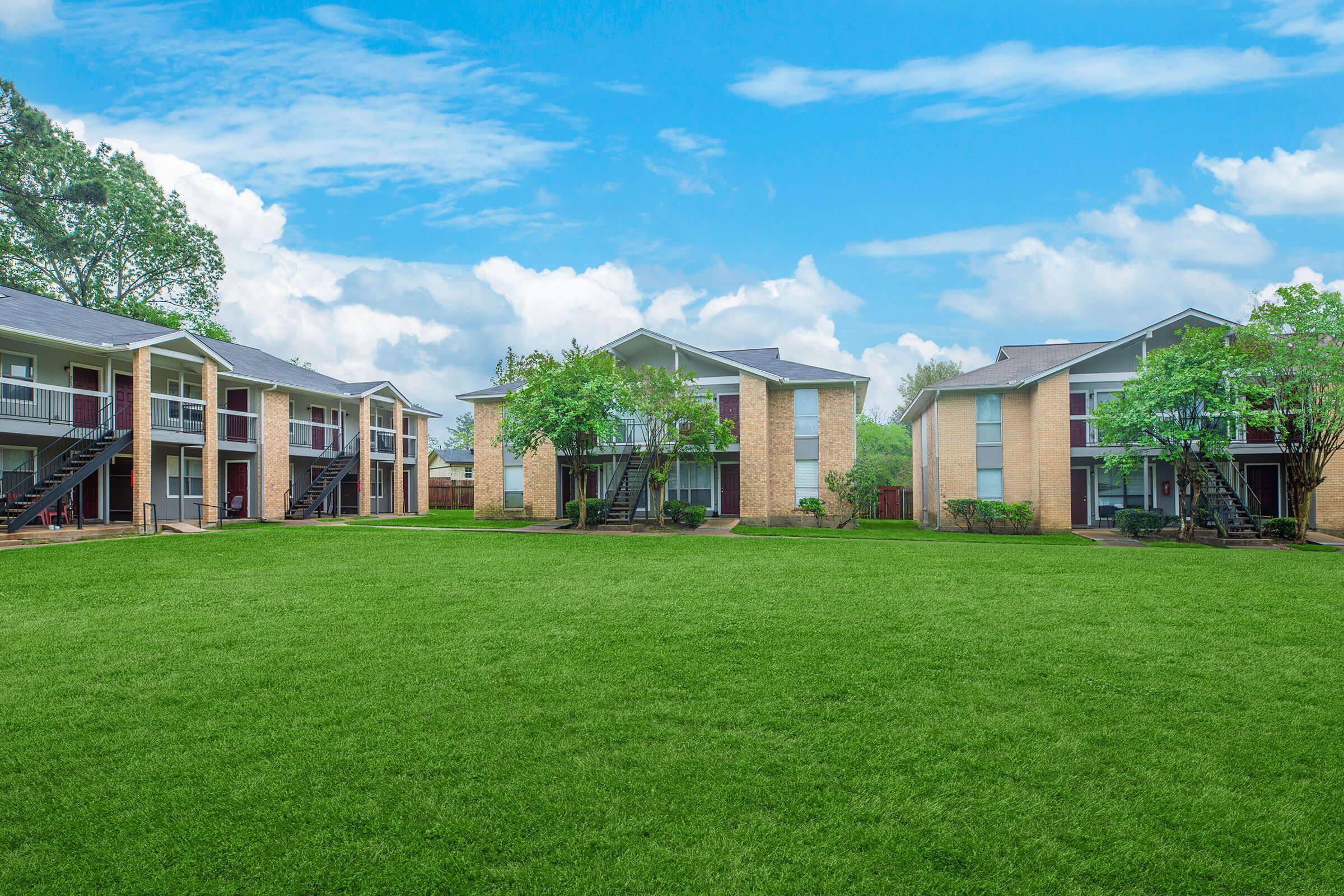 A serene apartment complex surrounded by lush green grass and trees, featuring two-story brick buildings with balconies. The sky is partly cloudy, providing a bright and inviting atmosphere.