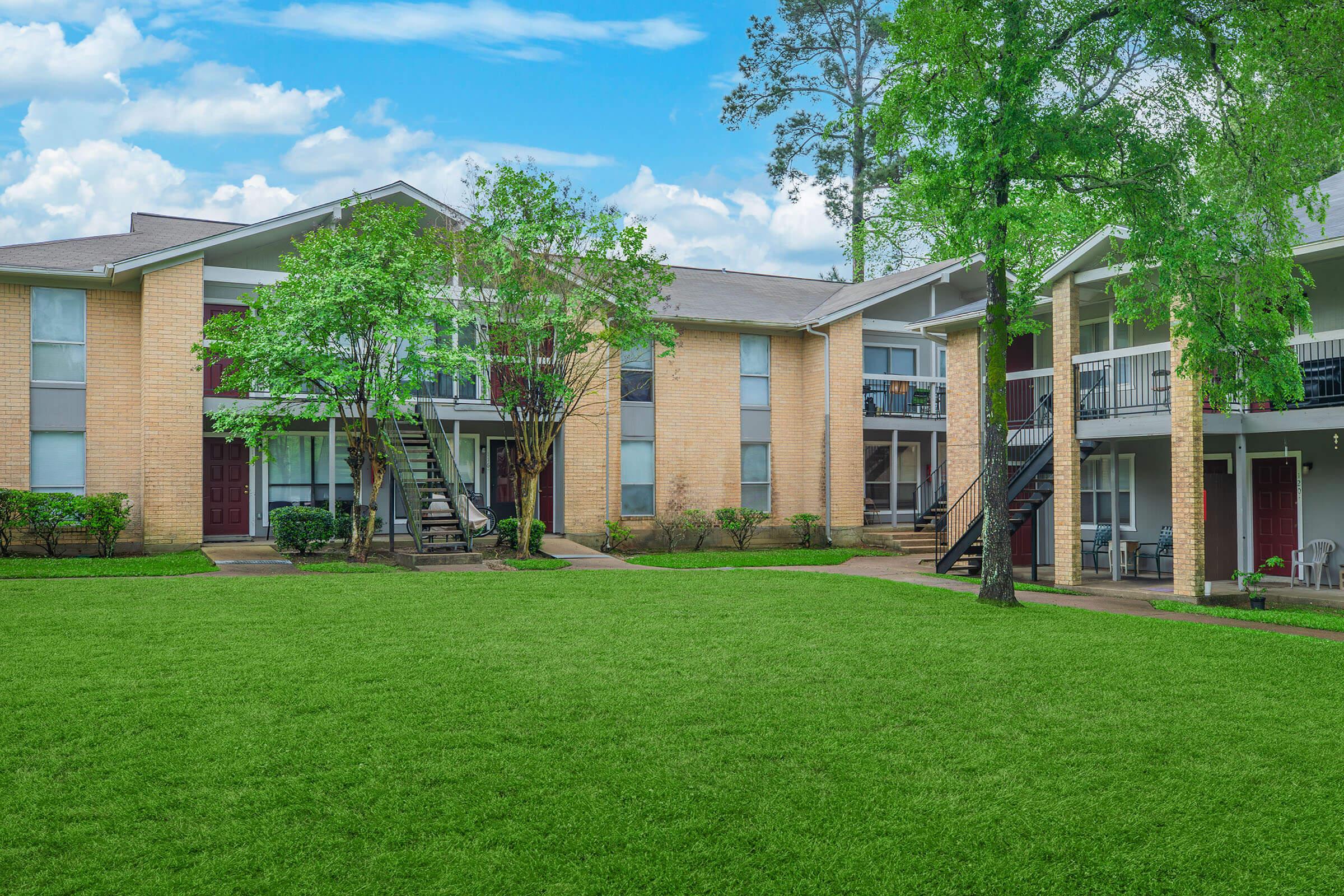 A well-maintained apartment complex featuring two-story buildings with brick facades, surrounded by lush green lawns and trees. The scene includes walking paths leading to the entrances, a clear blue sky with clouds, and neatly arranged balconies.